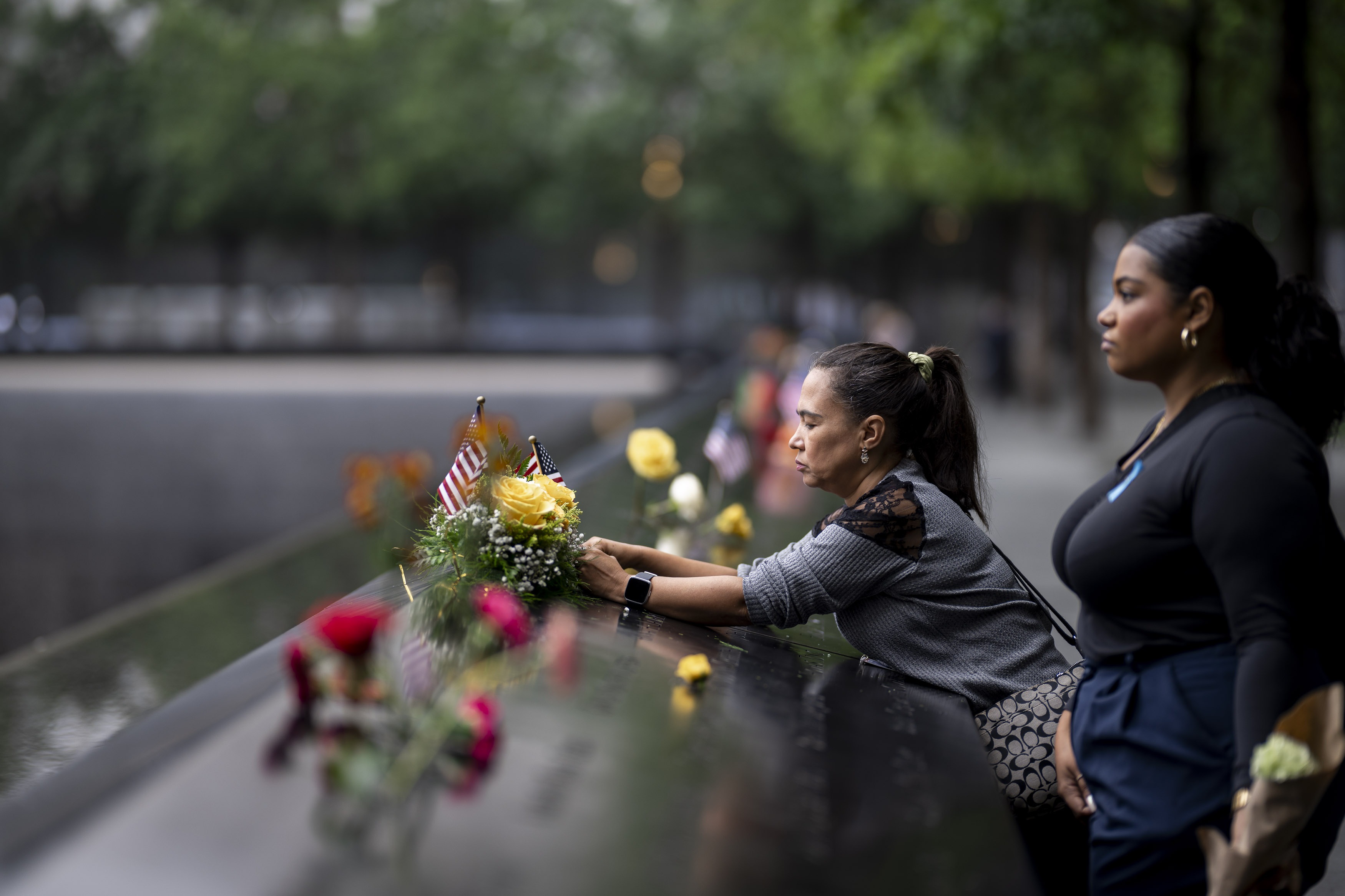 Two women place flowers and a small flag at the edge of a memorial reflecting pool.