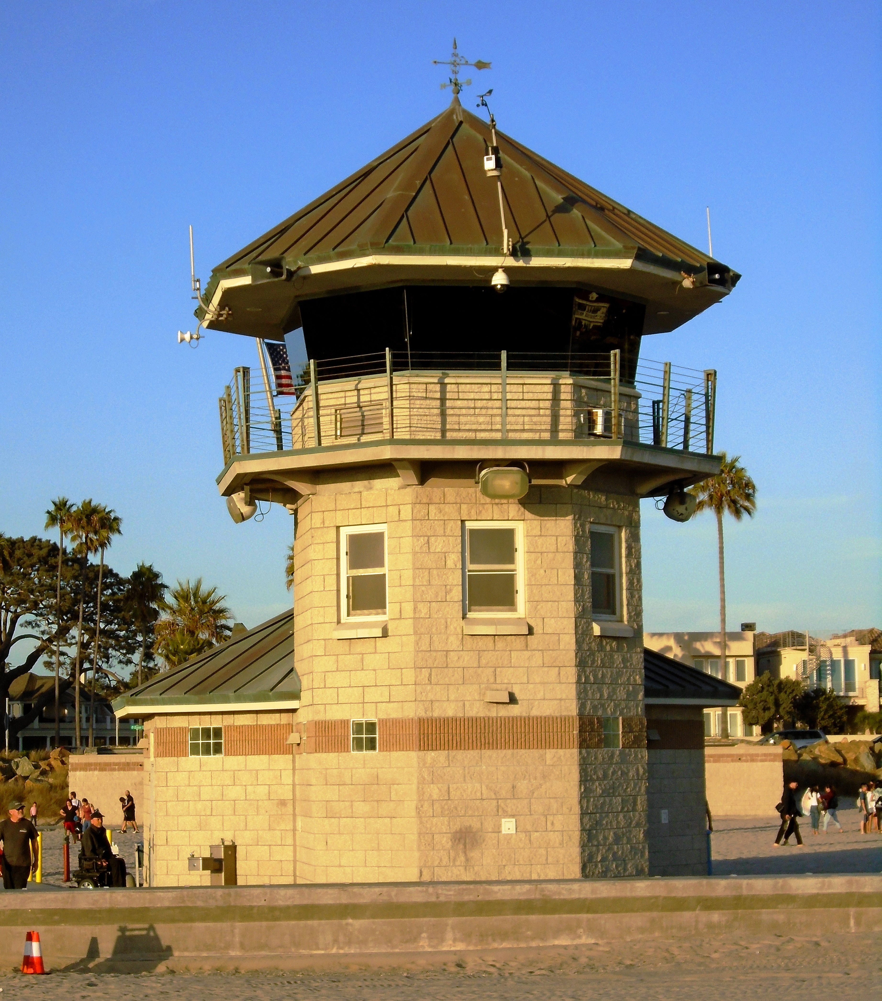 Octagonal stone-and-metal lifeguard tower on a sunny beach with palm trees nearby.