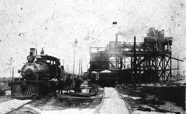 Black-and-white historical photo of workers and a steam locomotive at a phosphate loading facility.