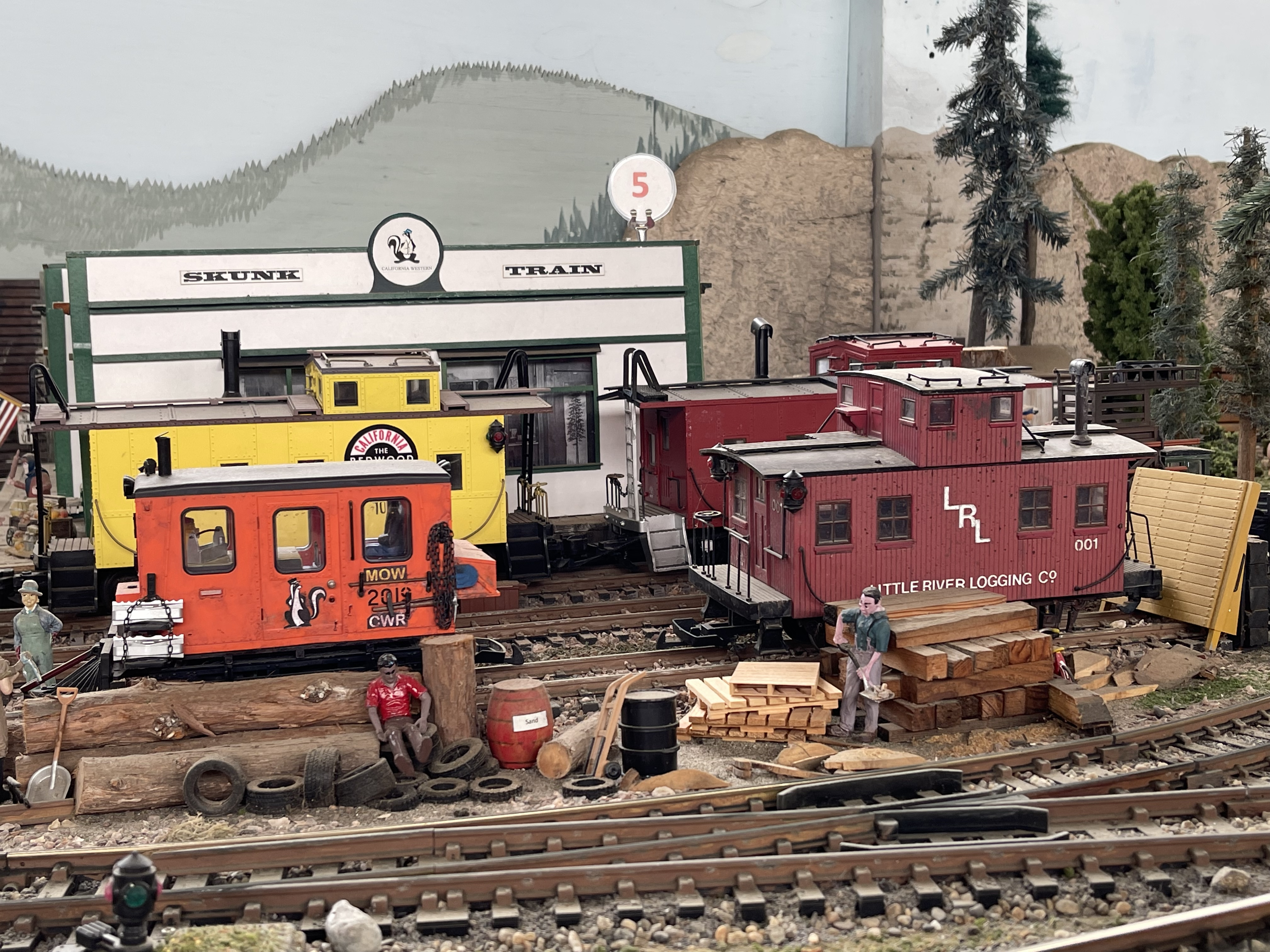 Model train layout with red and orange cabooses on tracks against a painted mountain backdrop.