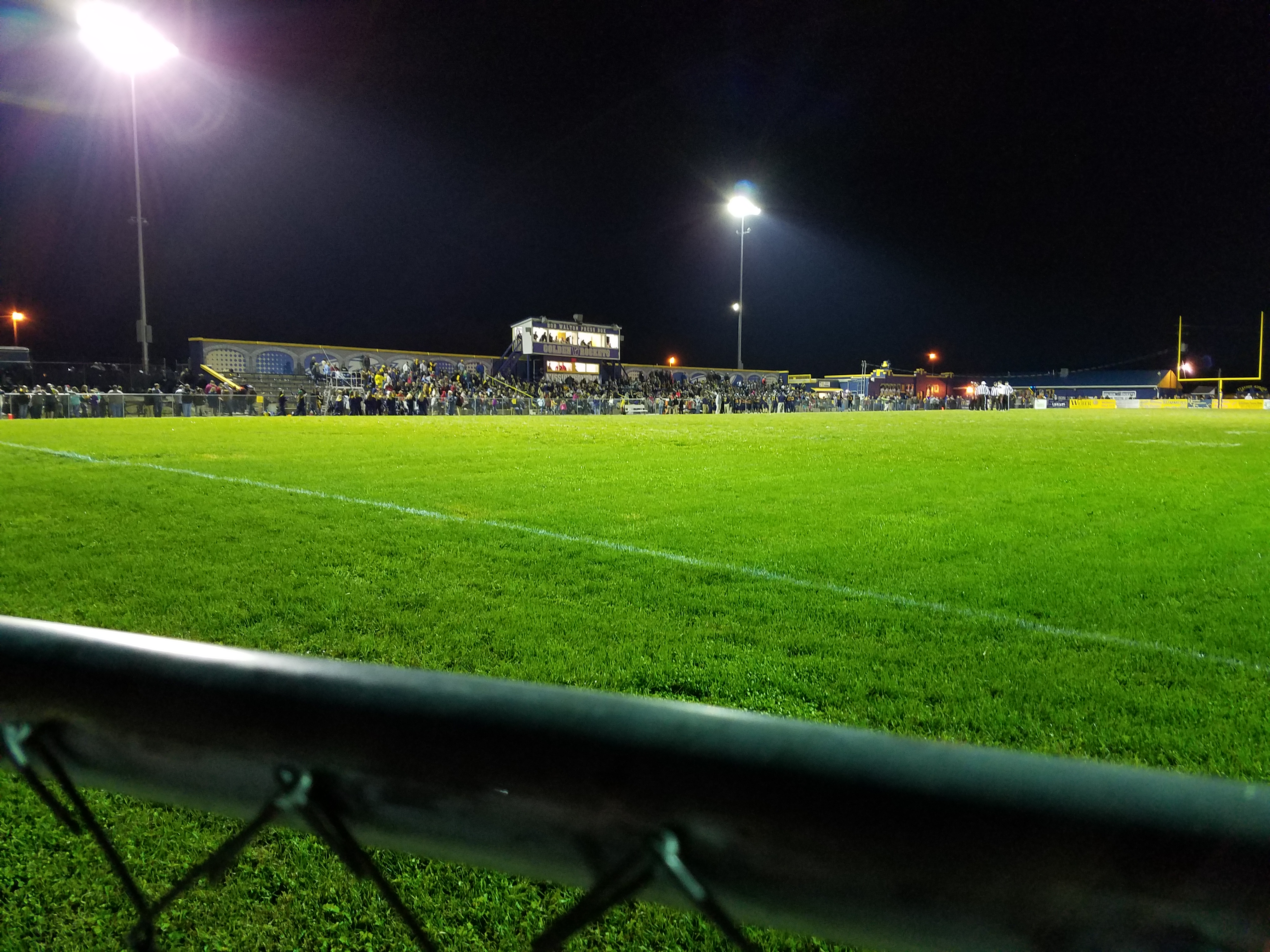 High school football stadium lit up at night with packed bleachers and a press box.