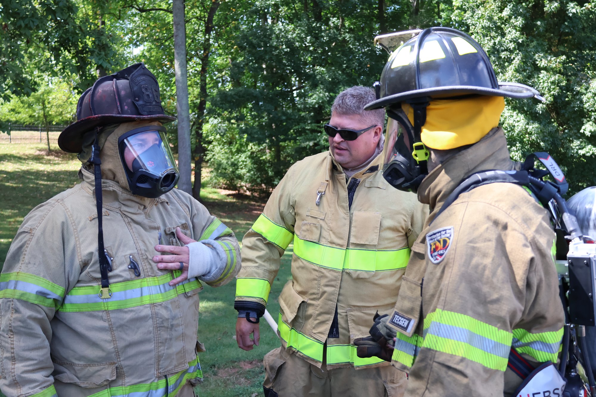 Three people in firefighter turnout gear and helmets converse outdoors in a wooded area during a training exercise.
