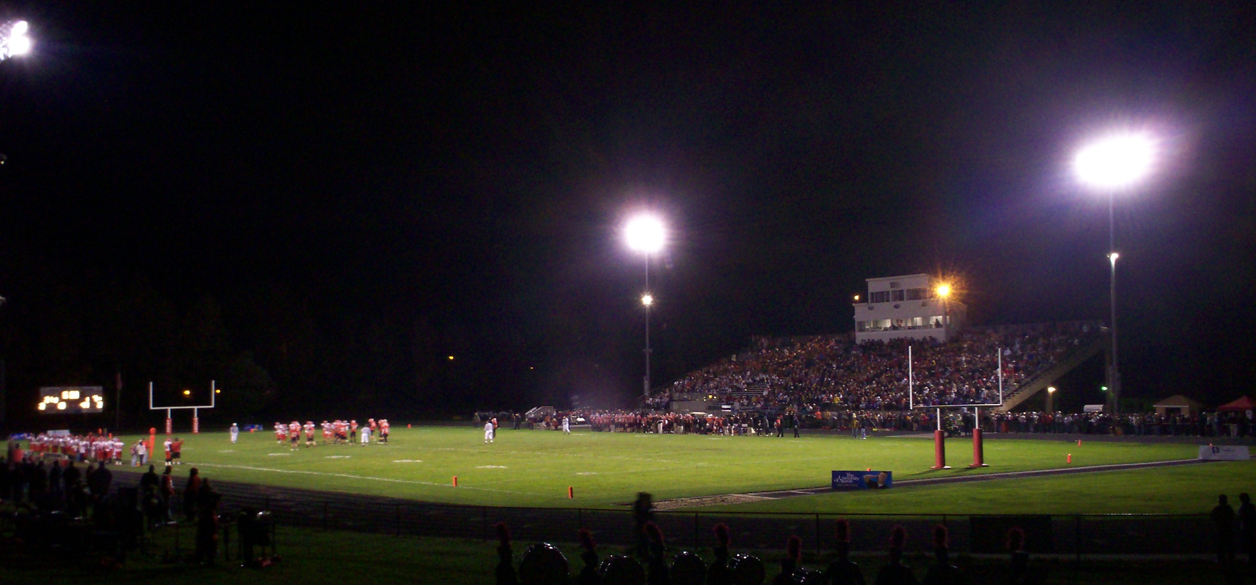 High school football stadium lit up at night with players on the field and a full grandstand.
