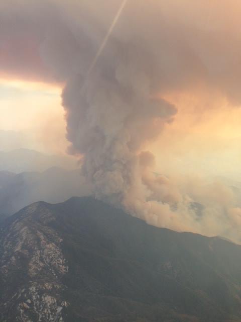 Aerial view of a towering dark smoke column rising from a wildfire on a mountain ridge.