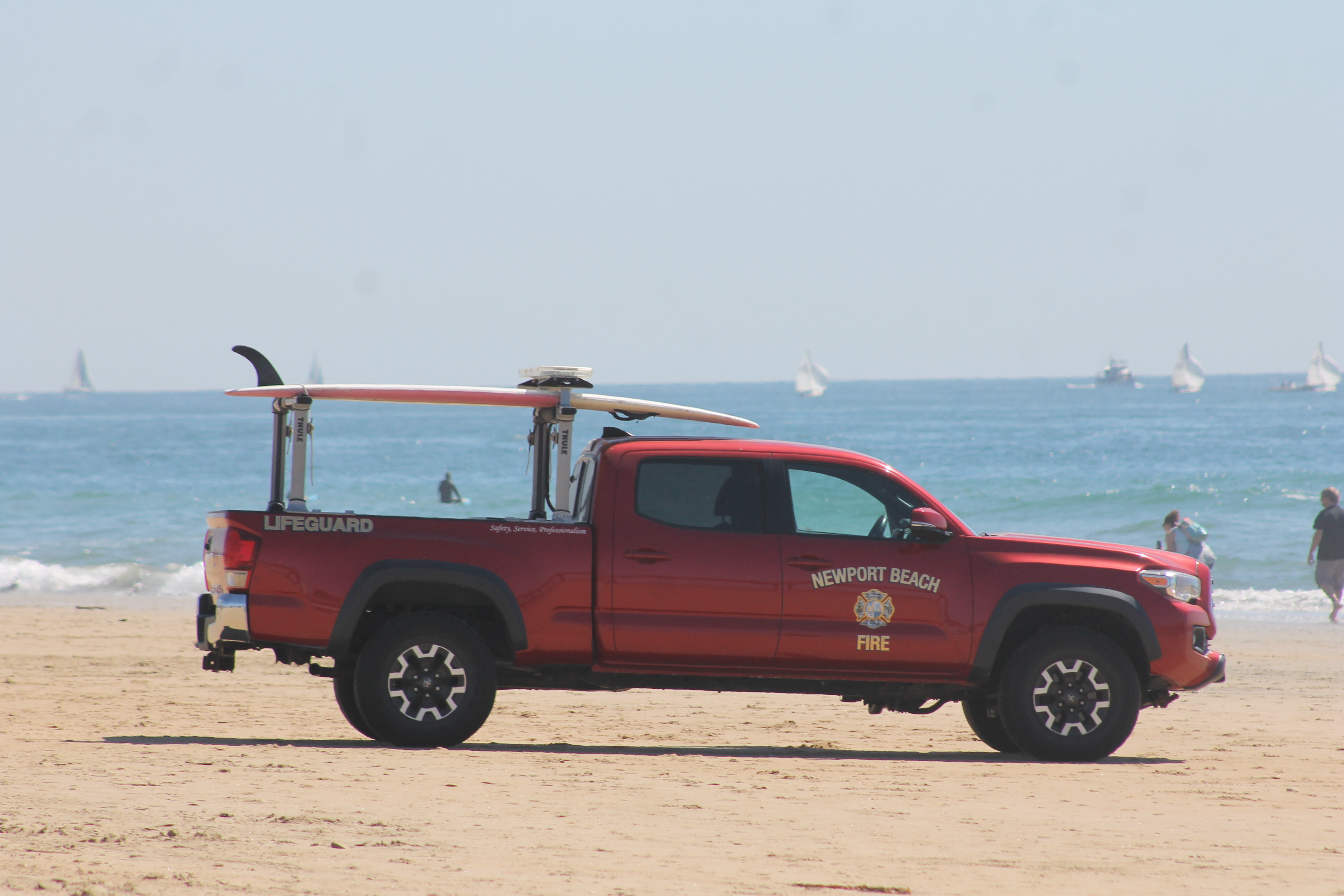 Red Newport Beach Fire Department lifeguard truck parked on a sandy beach with surfboards on the roof.