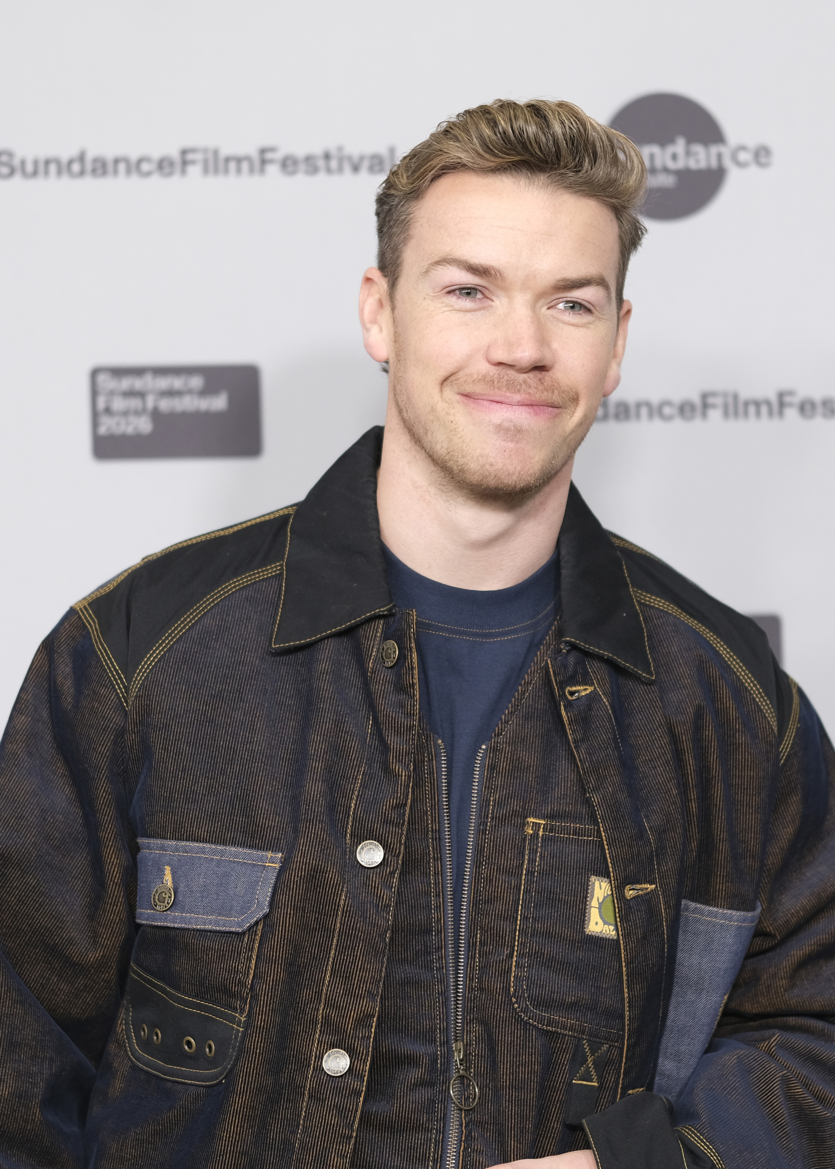 Man with short blond hair in a dark denim jacket standing before a Sundance Film Festival backdrop.