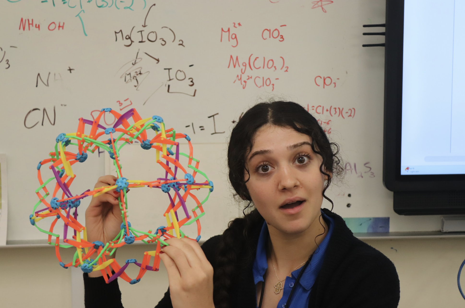 A woman holds a colorful molecular model in front of a whiteboard covered in chemistry formulas.
