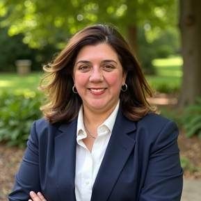 Woman in navy blazer smiling with arms crossed outdoors in front of green trees.