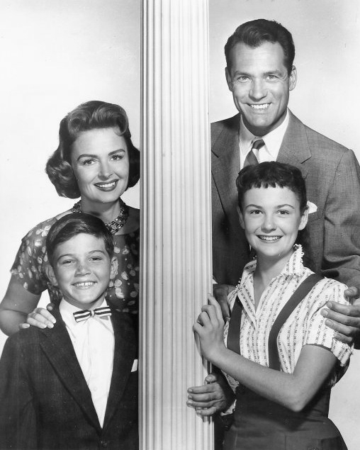Black-and-white cast photo of four people posed around a white decorative column.
