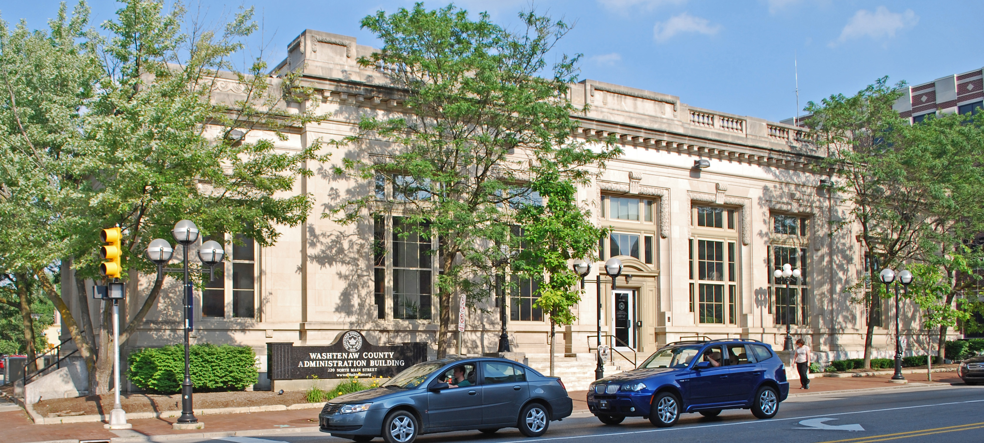 Neoclassical stone government building on a street corner with trees and parked cars.