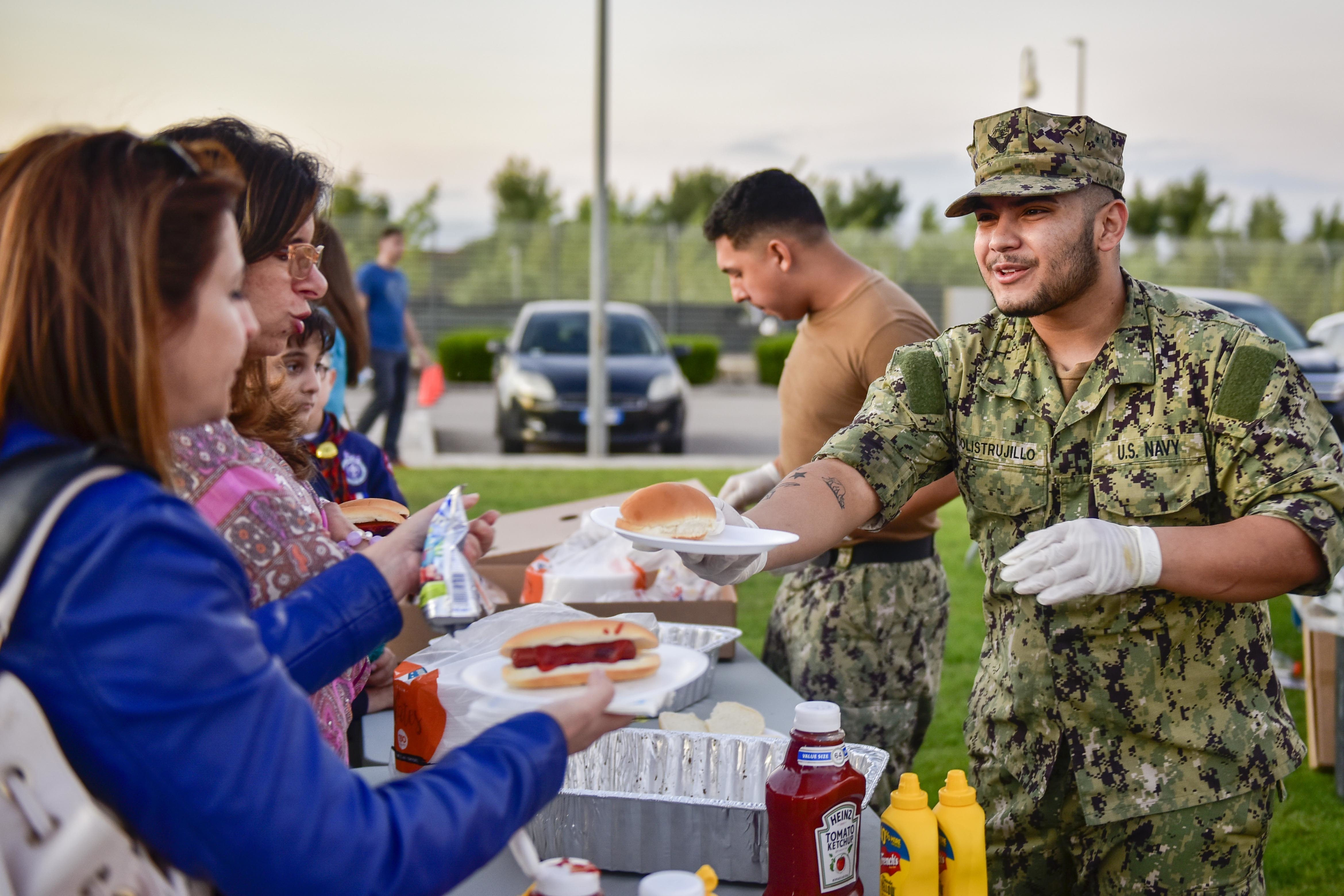 Person in camouflage uniform serving hamburgers to attendees at an outdoor community event.