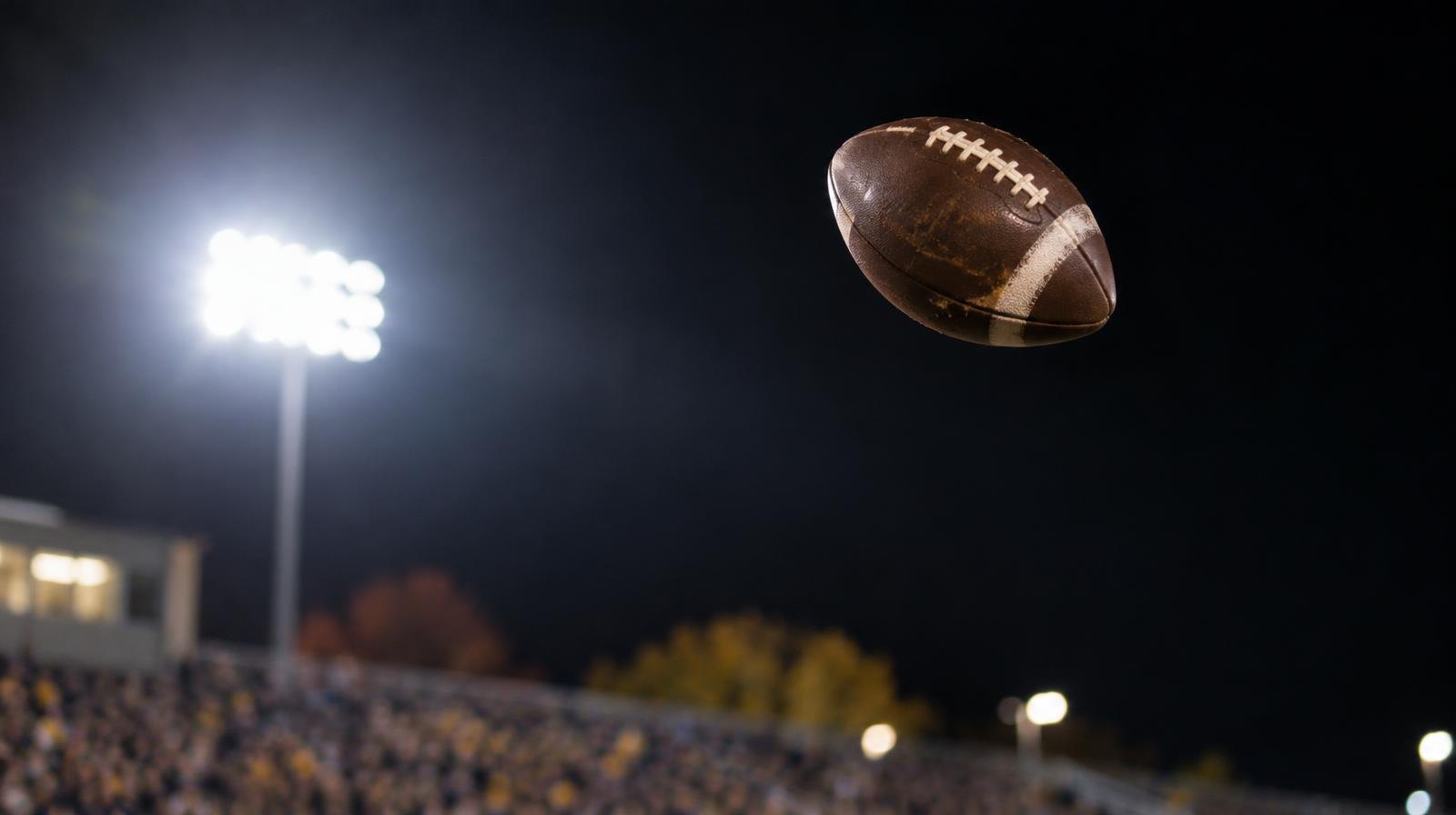 A football spiraling through the air against a dark night sky lit by stadium lights.