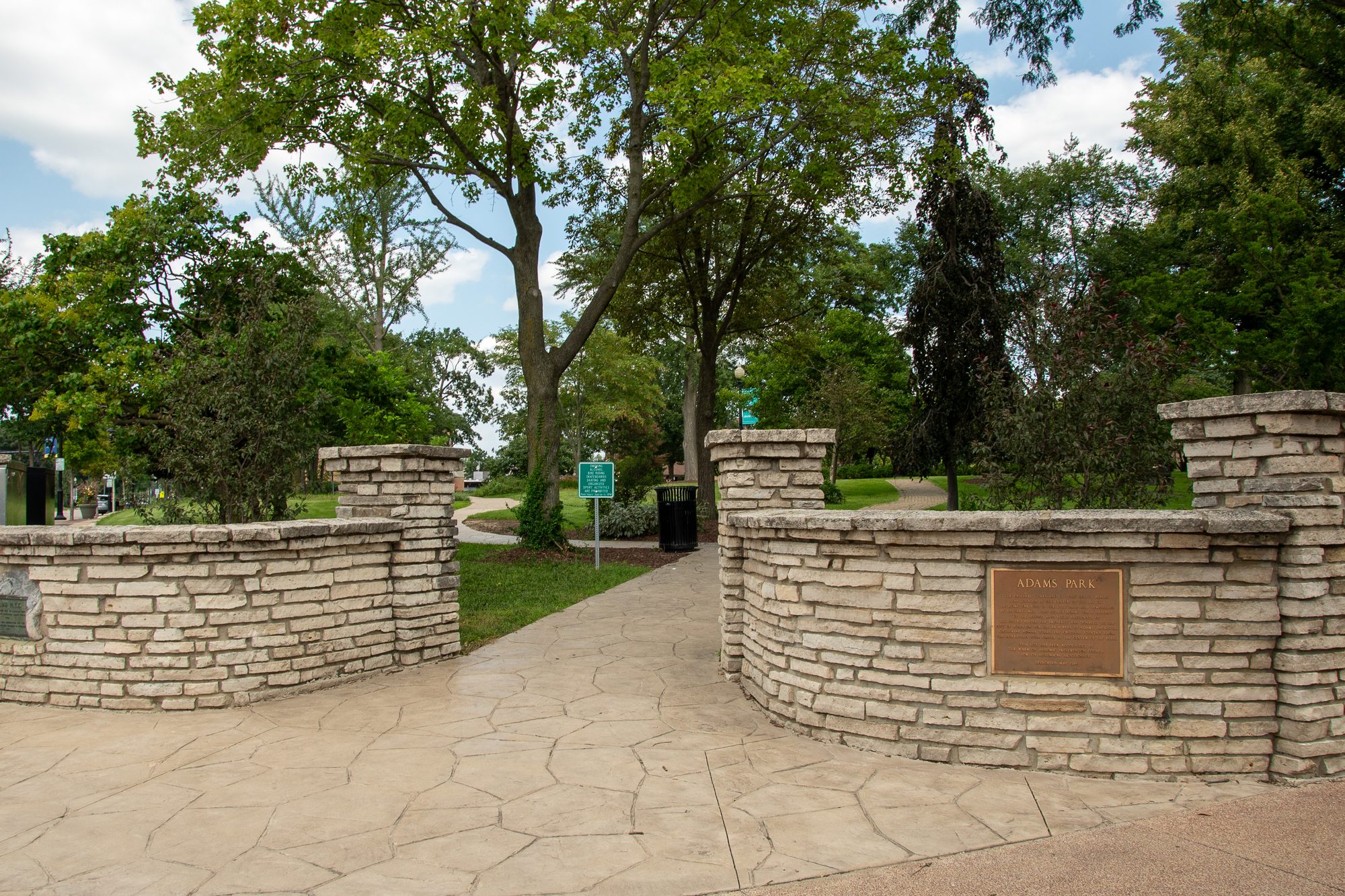 Stone entrance gate with curved limestone walls leading into a tree-lined park pathway.