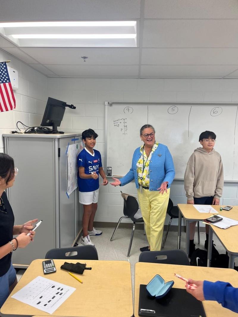 An adult woman in a blue cardigan interacts with a student at a classroom whiteboard.