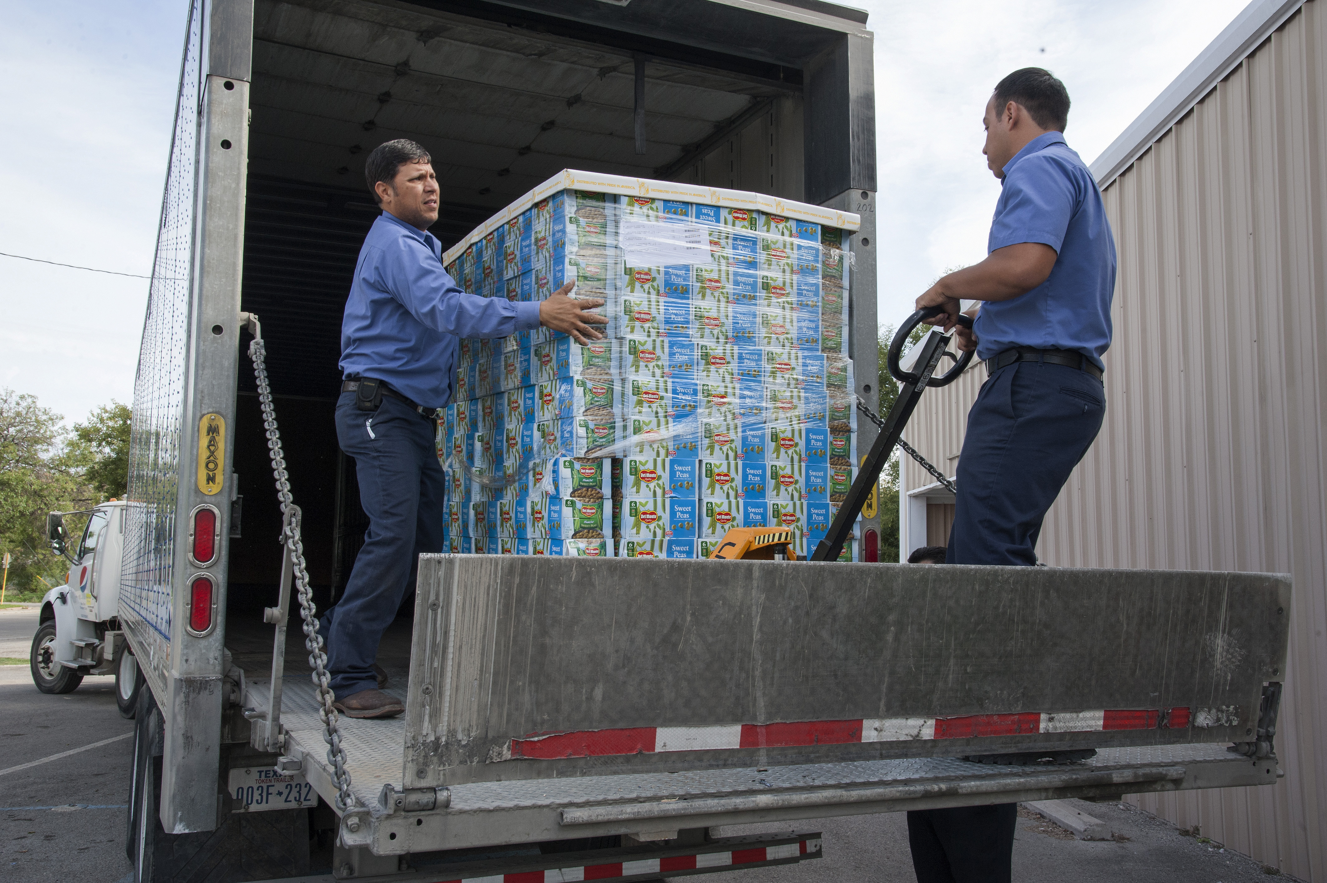 Two workers in blue uniforms unloading a pallet of boxed food from a delivery truck.