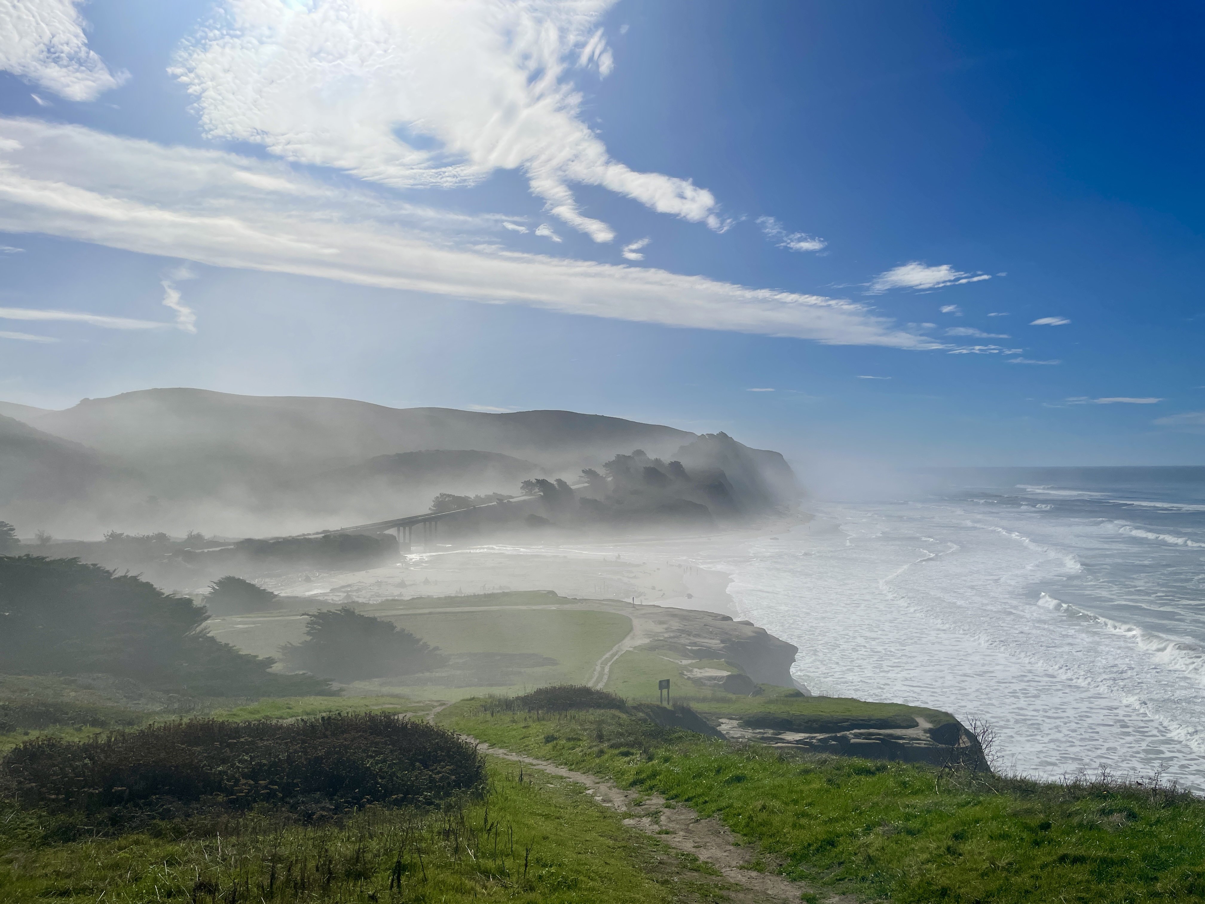 Clifftop view of a coastal lagoon, sandy beach, and misty bluffs with a highway in the distance.
