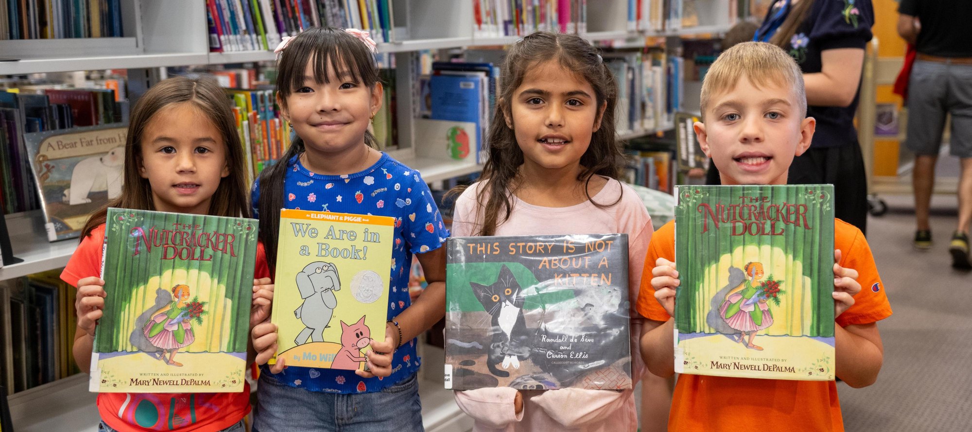 Four young children stand in a library, each holding a book and smiling.