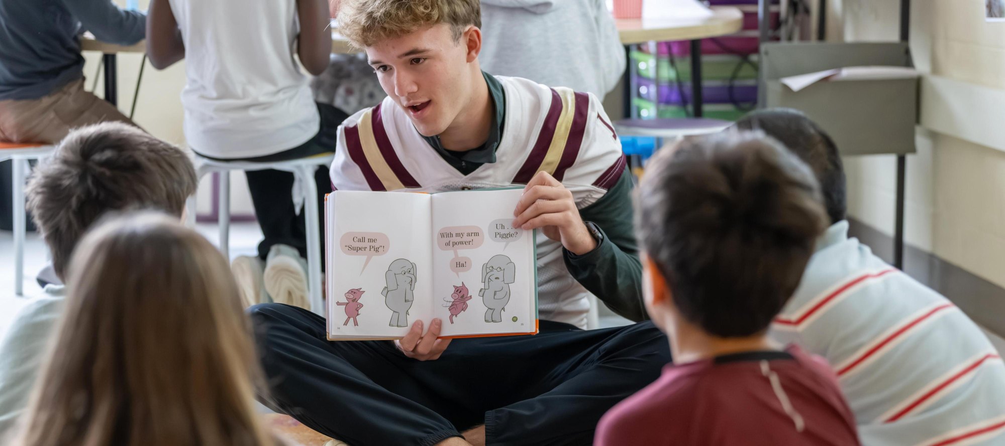A teenager in a football jersey reads a picture book to young children seated on a floor.