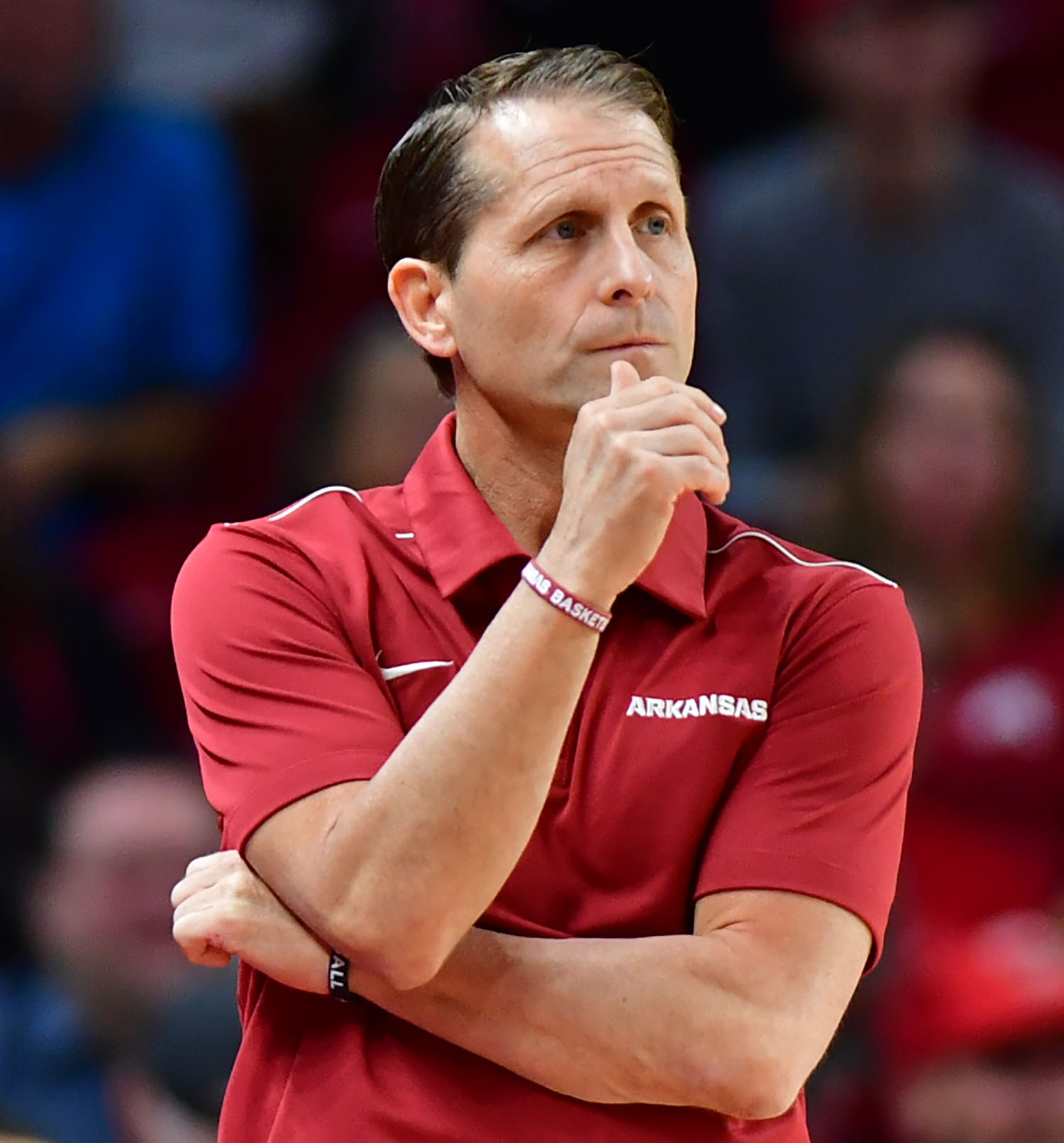 Man in a red polo shirt stands on a basketball sideline with arms crossed.