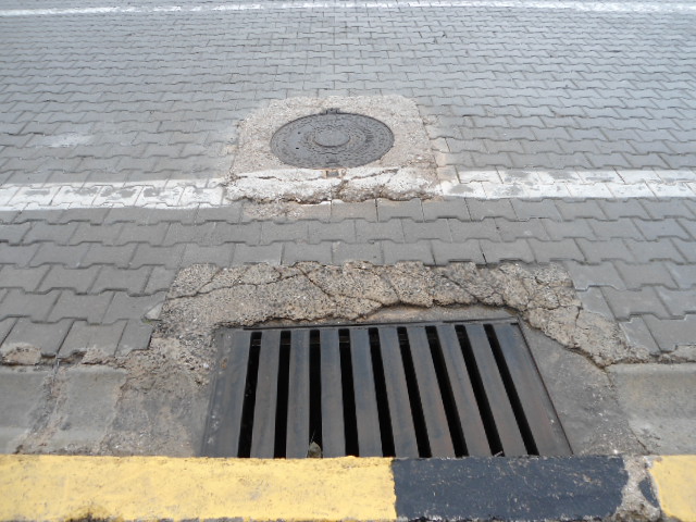 A rectangular metal storm drain grate set in cracked asphalt at a curb.