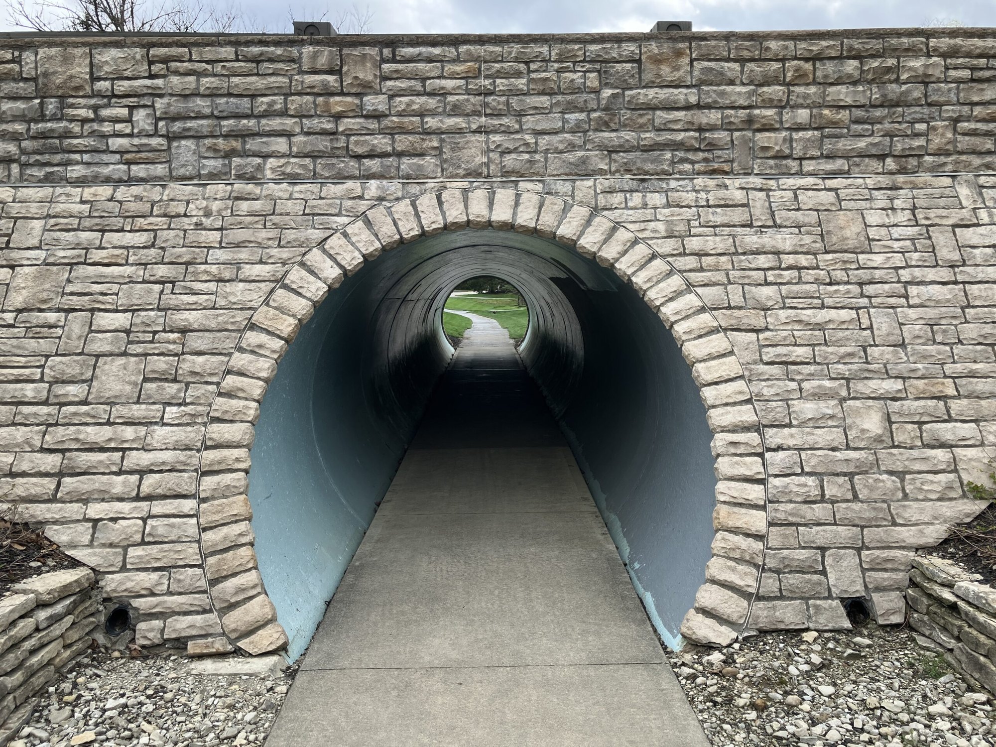 Stone arch entrance to a cylindrical concrete pedestrian tunnel with a paved walkway inside.