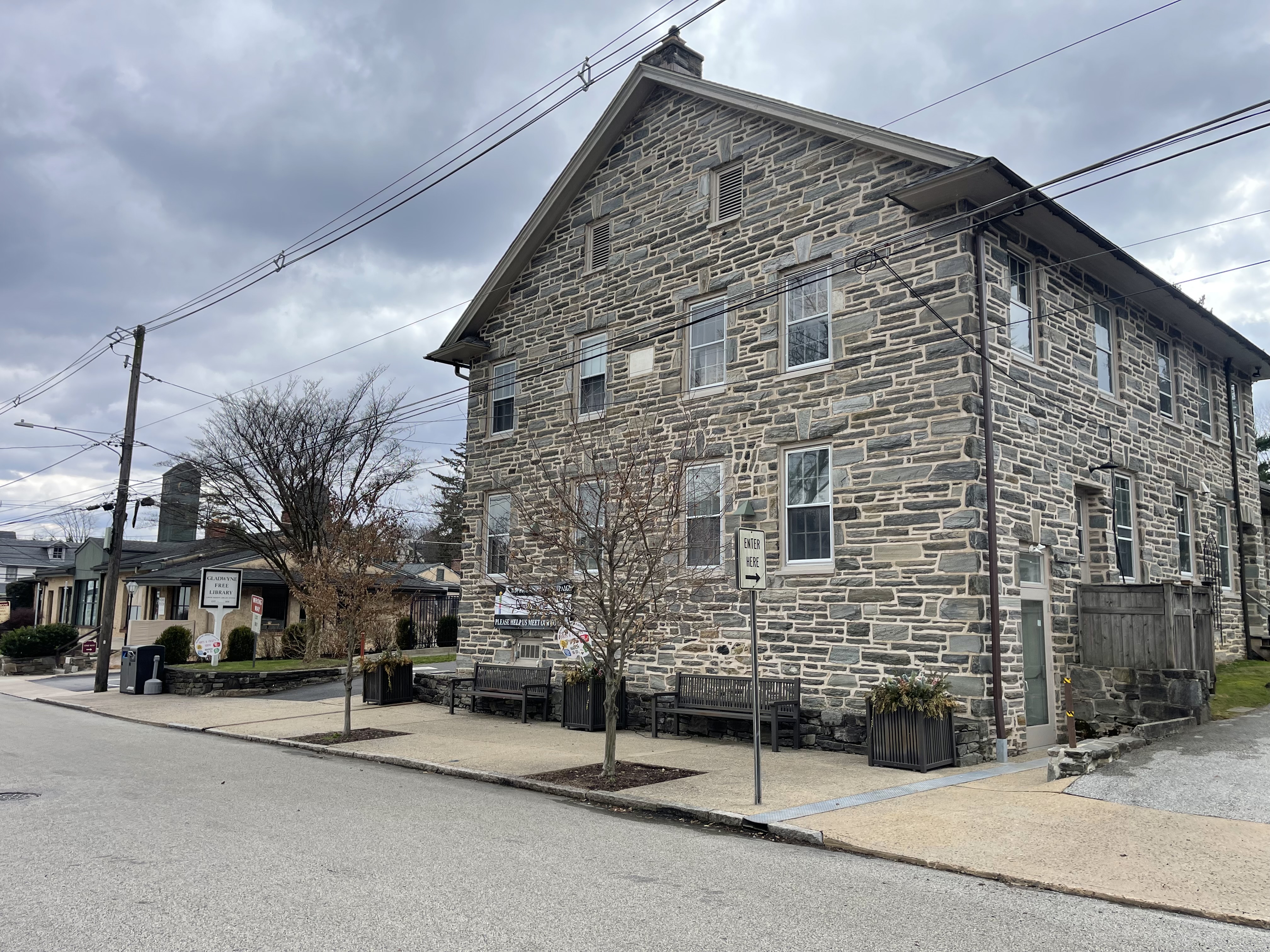 Three-story grey stone library building on a street corner with sidewalk benches and bare trees.