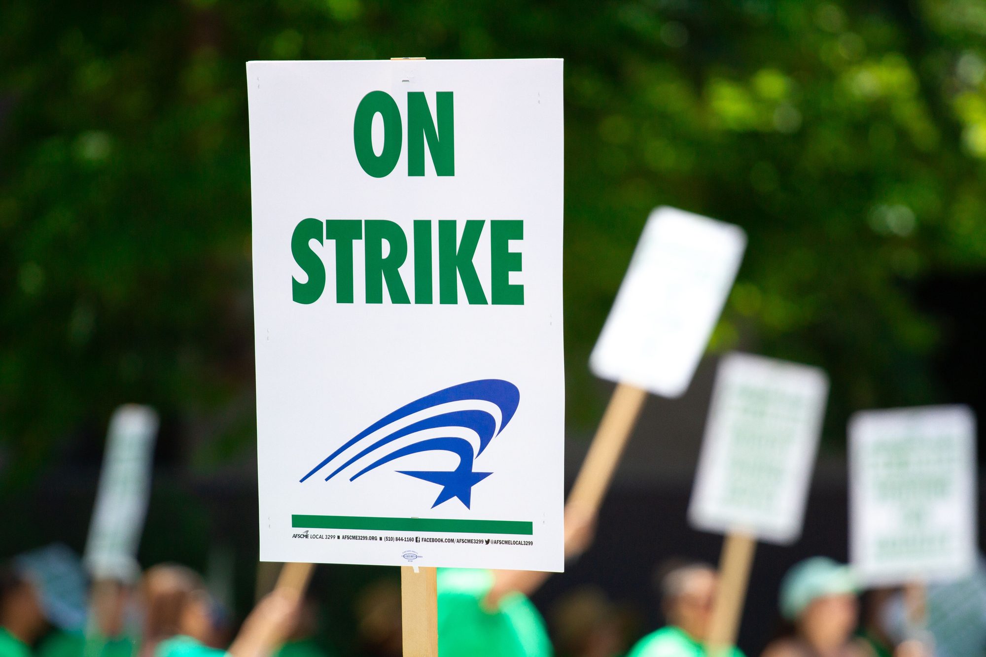 A white sign reading "ON STRIKE" with a union logo, held above a crowd of protesters holding blank picket signs.