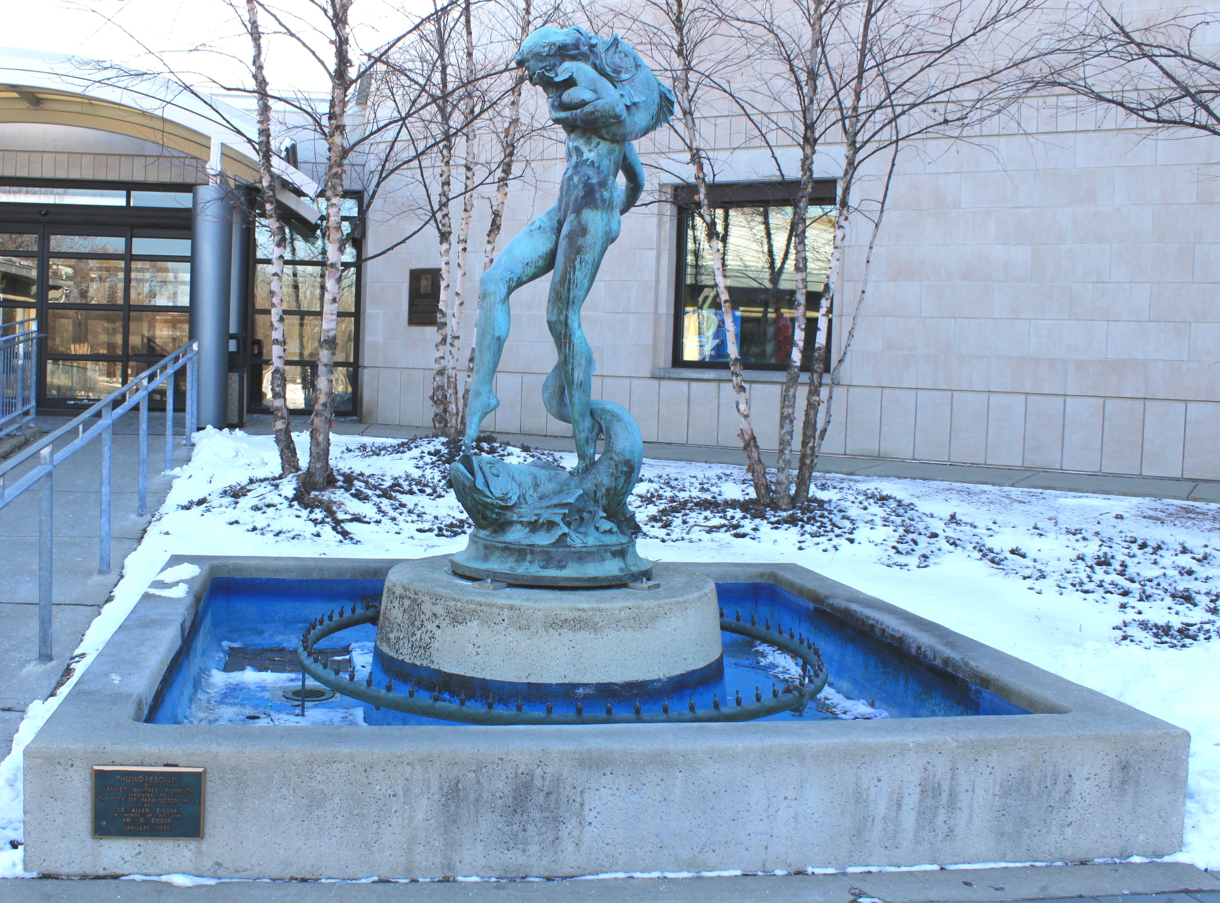 Bronze fountain sculpture on a pedestal outside a library building in winter with snow on the ground.