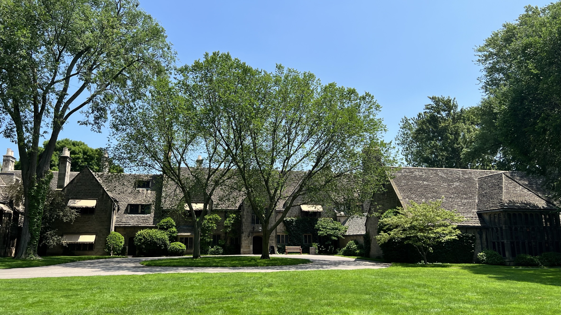 Stone manor house with steeply pitched roofs surrounded by mature trees and a green lawn.
