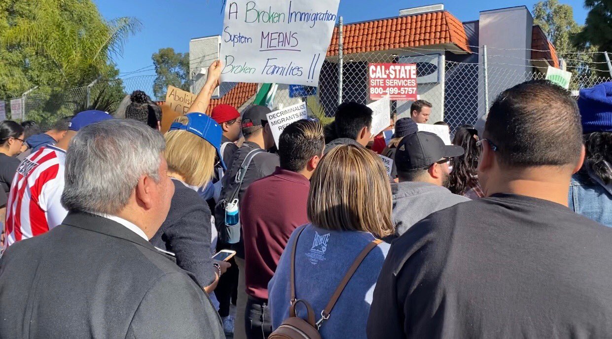Crowd of protesters at an outdoor rally, one holding a sign about immigration and broken families.