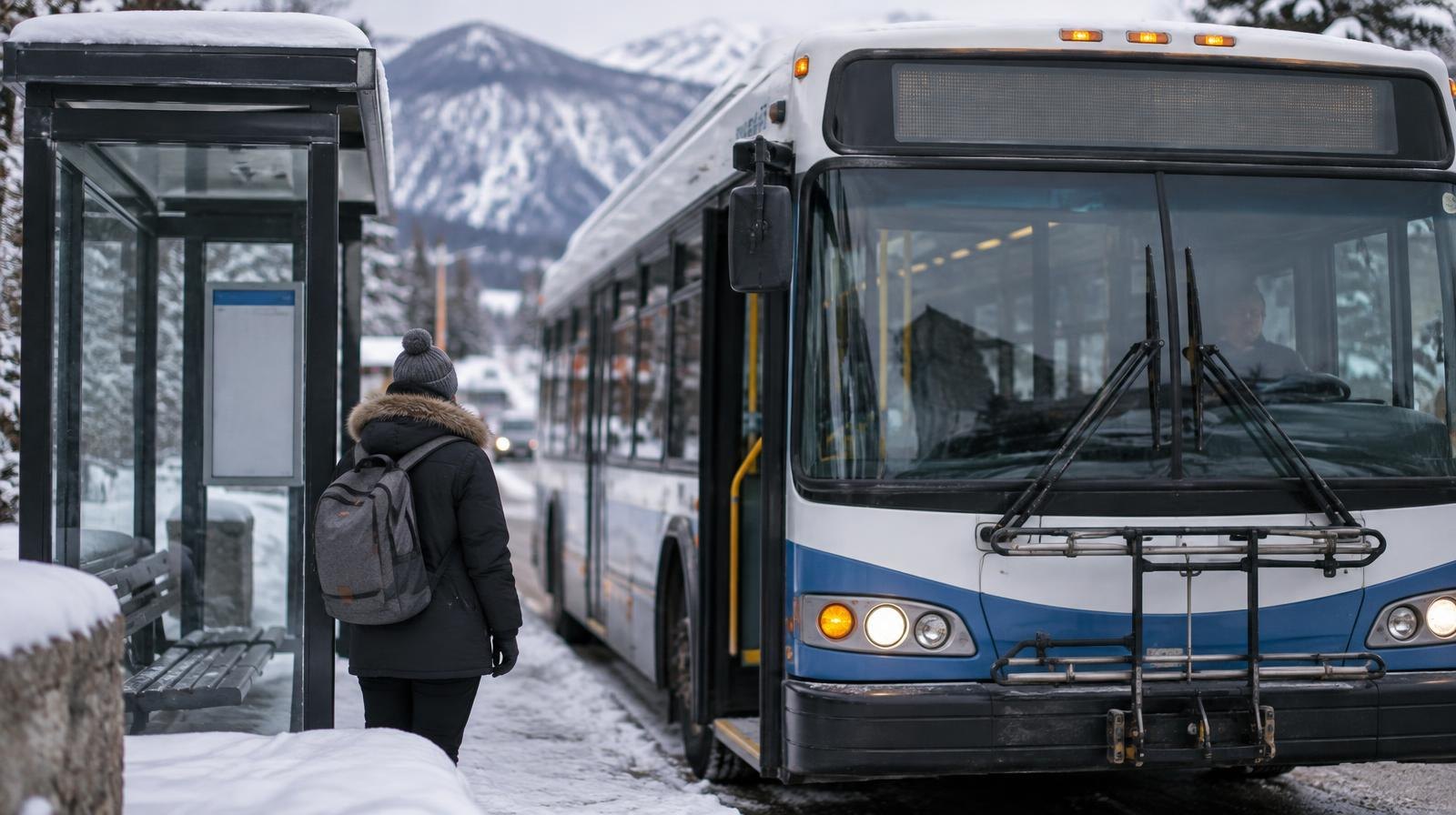 A transit bus stopped at a snow-covered shelter with mountains in the background.