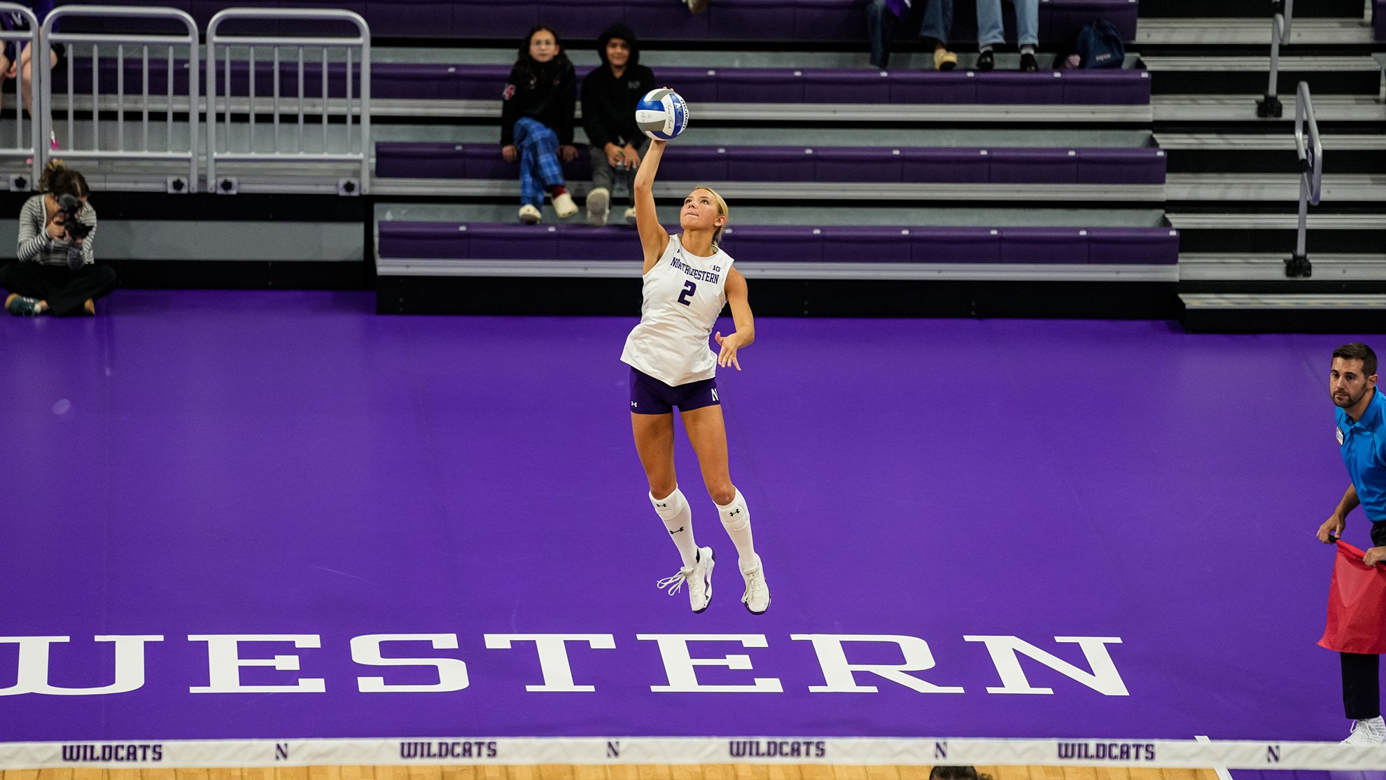 A volleyball player in a white uniform jumps to serve on a purple indoor court.