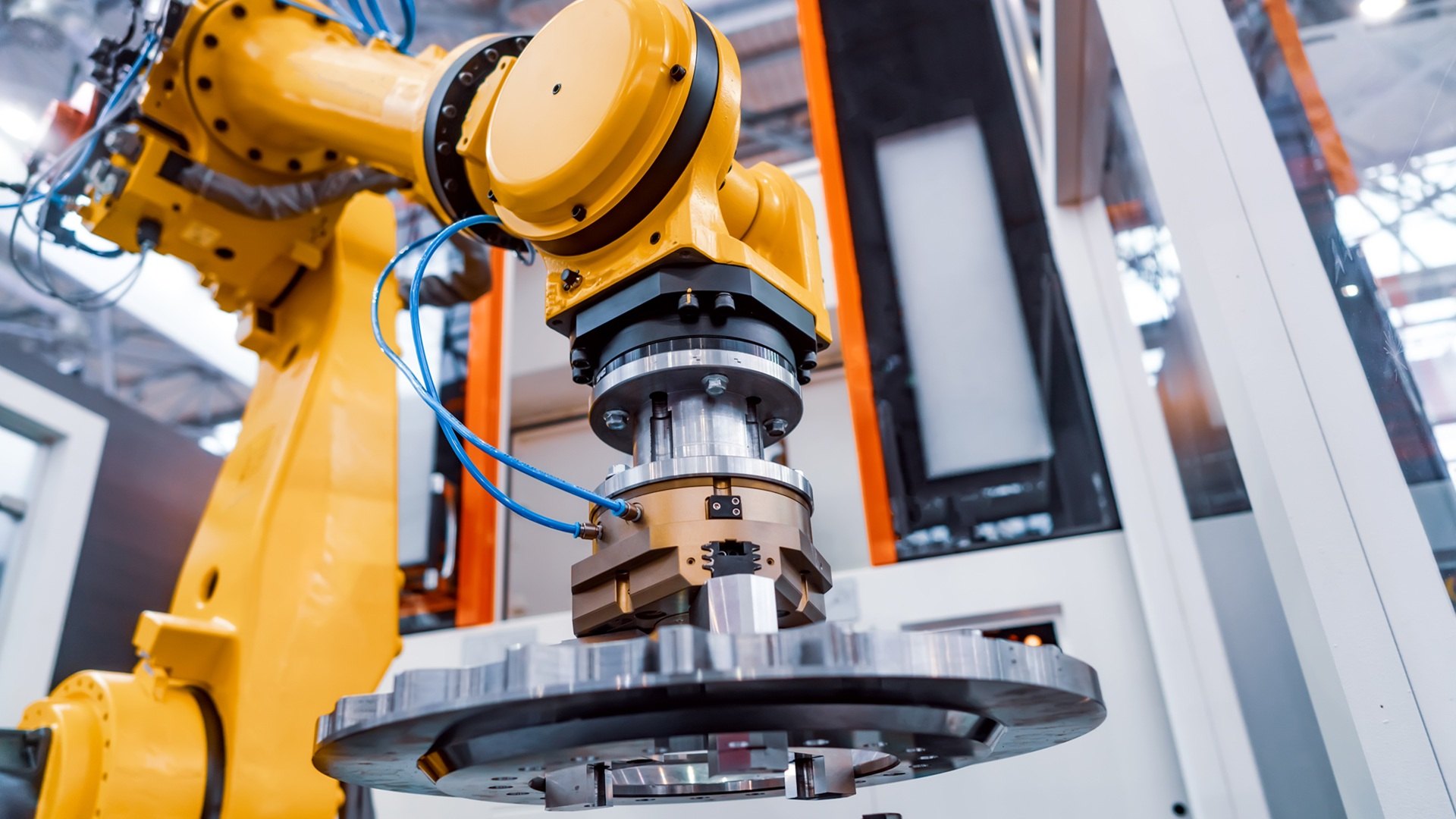 Yellow industrial robotic arm gripping a large circular metal part on a factory floor.