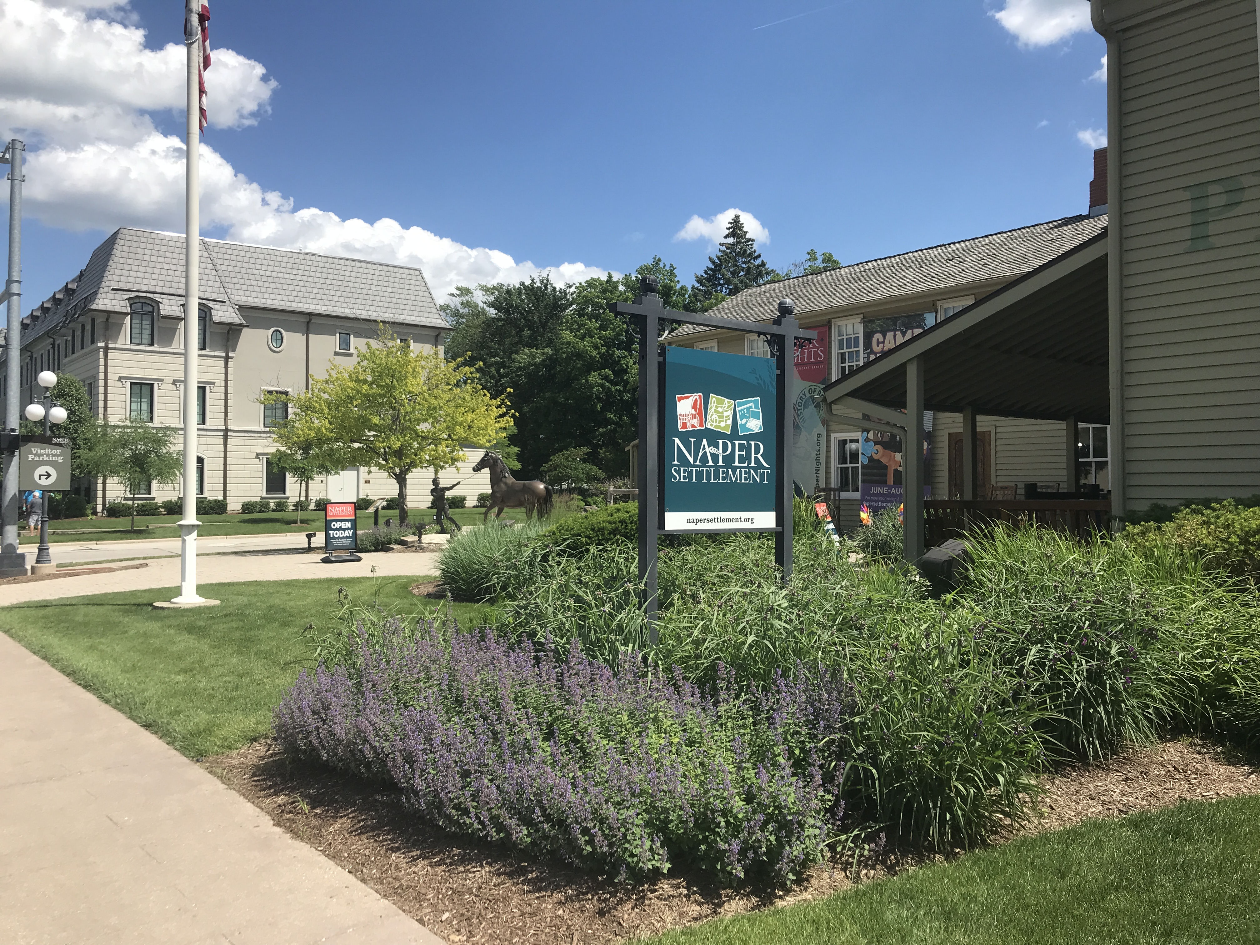 Sign reading Naper Settlement on a post with flowering plants and buildings behind.