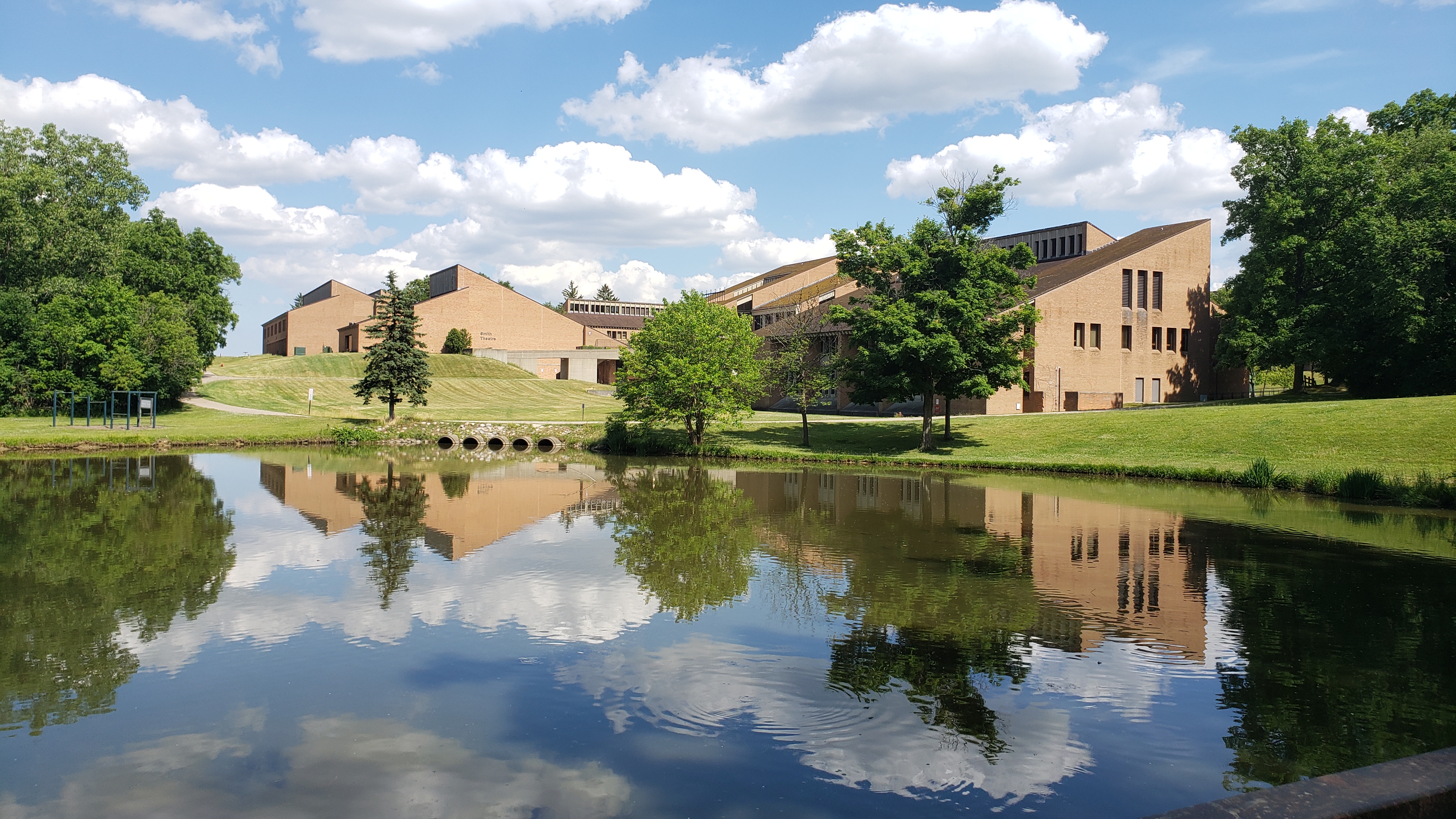 Brick college buildings reflected in a calm campus pond under a partly cloudy sky.