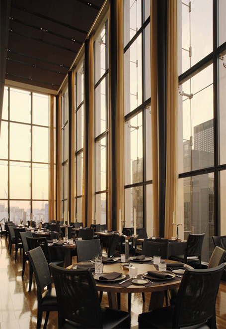 Upscale restaurant dining room with tall windows, set tables, and a city skyline at dusk.