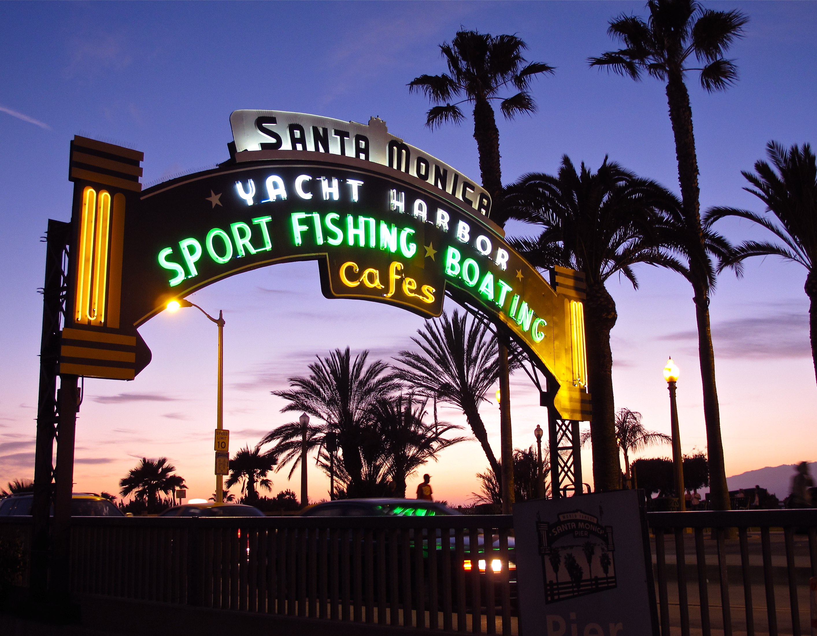 Illuminated neon arch sign at a pier entrance glowing against a purple twilight sky with palm trees.