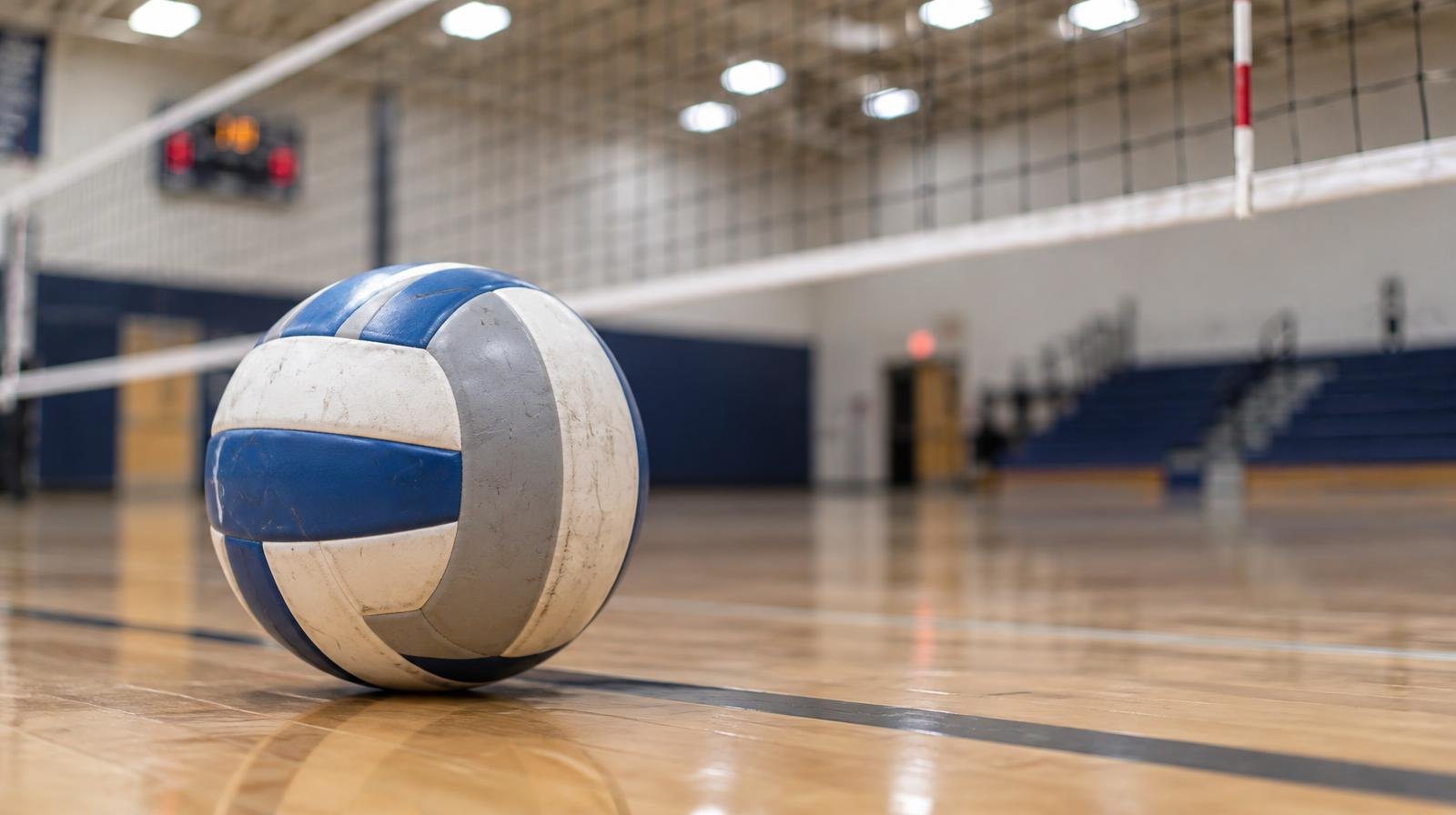 A volleyball resting on a hardwood gym floor near a volleyball net.