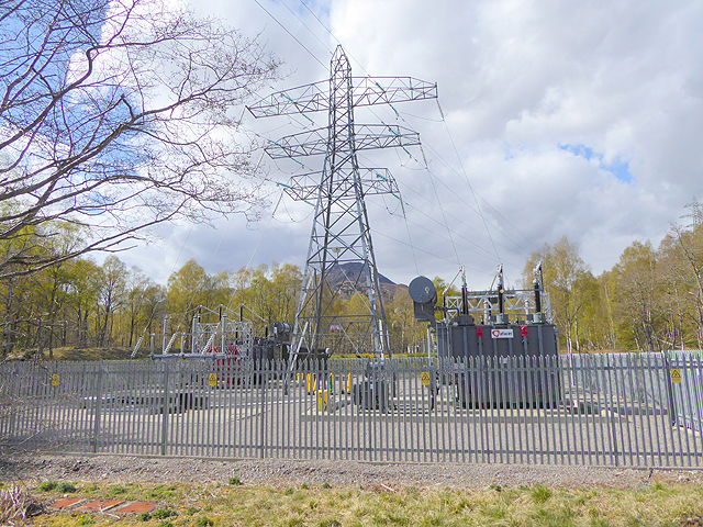 Fenced substation with large transformers and a tall steel lattice transmission tower.