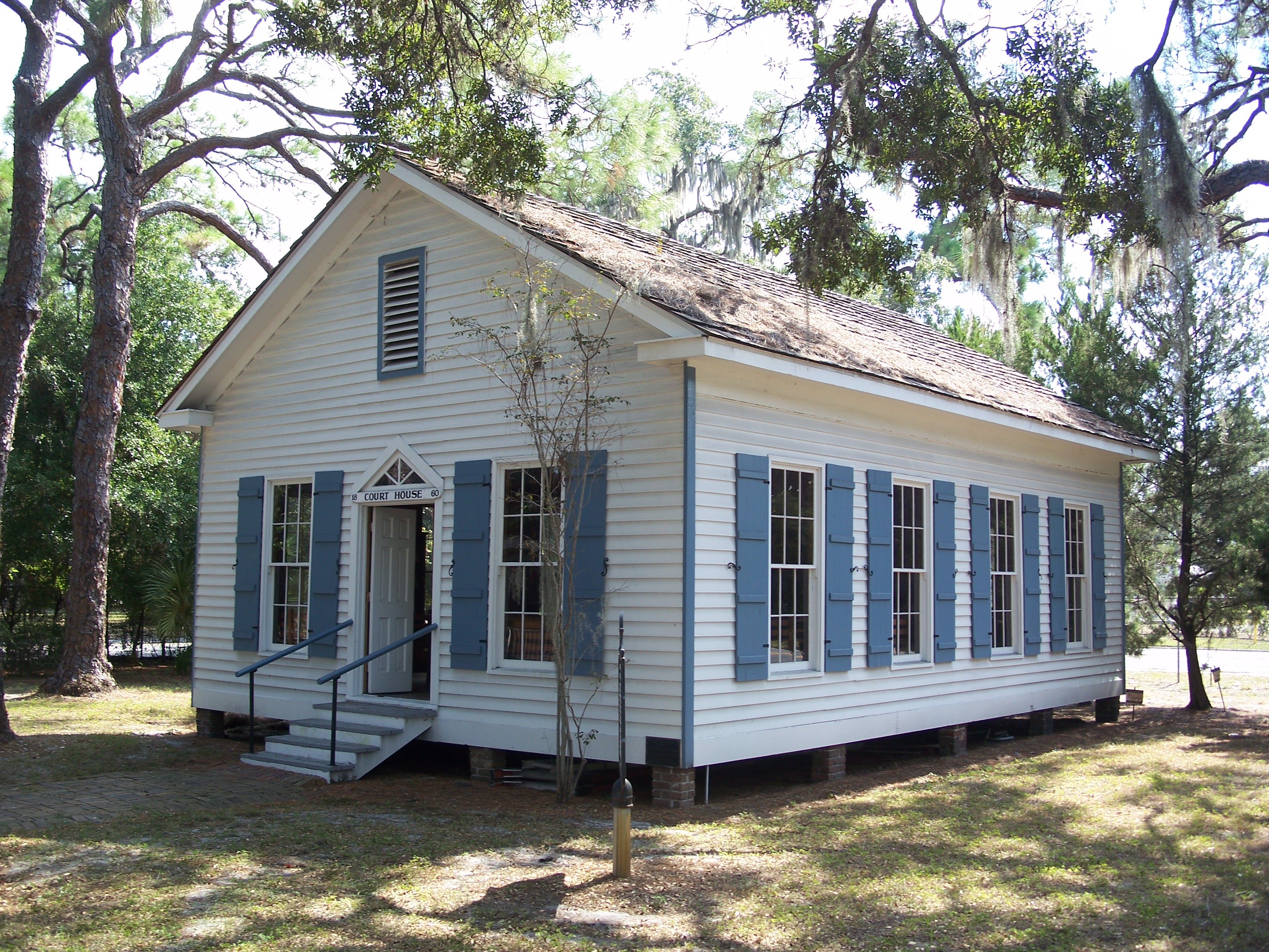 Small white historic wood-frame courthouse building with blue shutters, elevated on piers, surrounded by trees with Spanish moss
