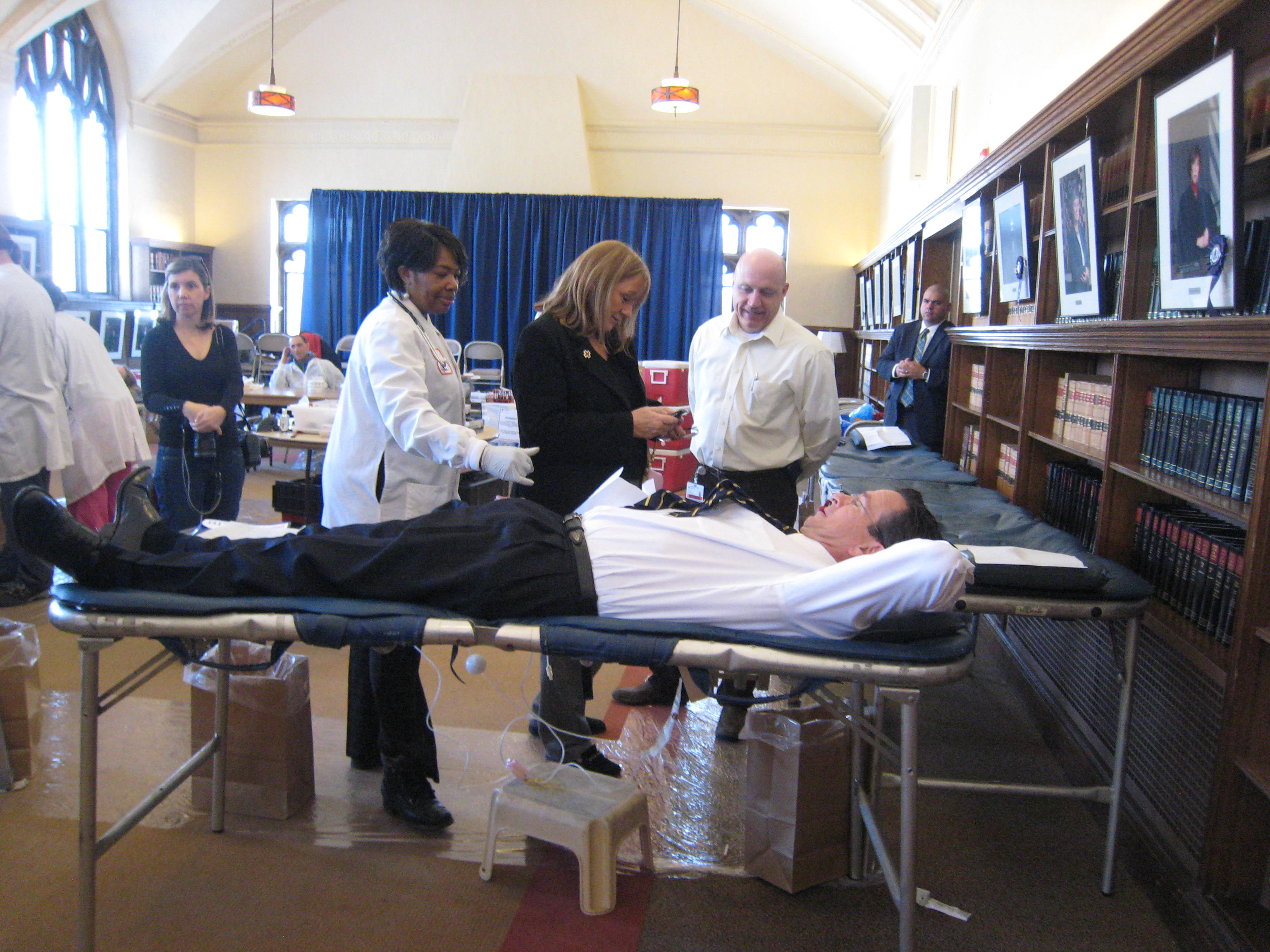 Man reclining on a donation cot at an indoor blood drive, attended by two medical staff members.