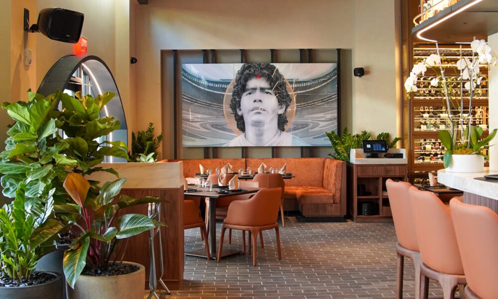 Upscale restaurant interior with leather booths, potted plants, wine display, and a large framed portrait of Diego Maradona.