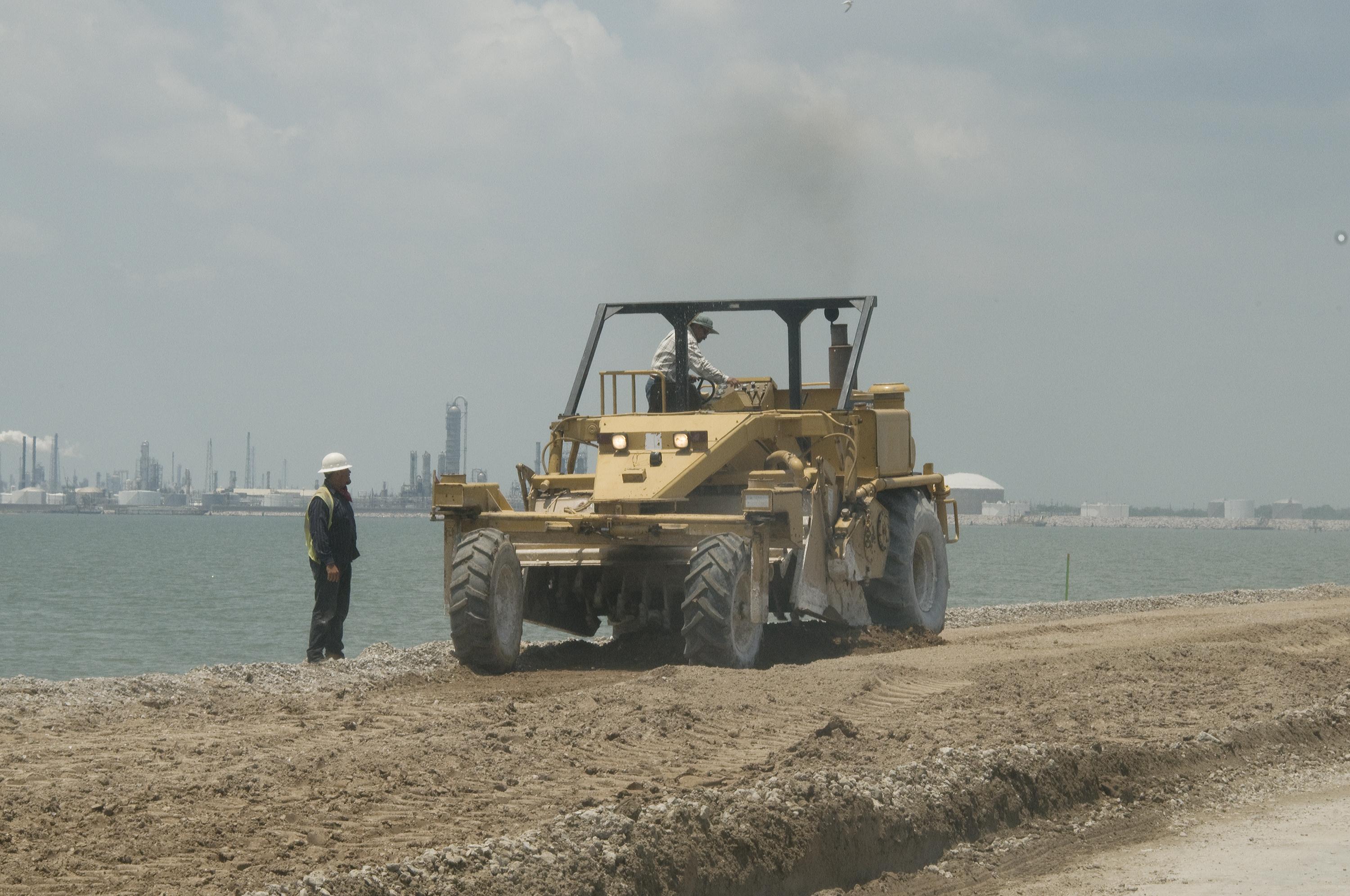 Large yellow road compactor grading a dirt surface, worker in hard hat standing nearby.