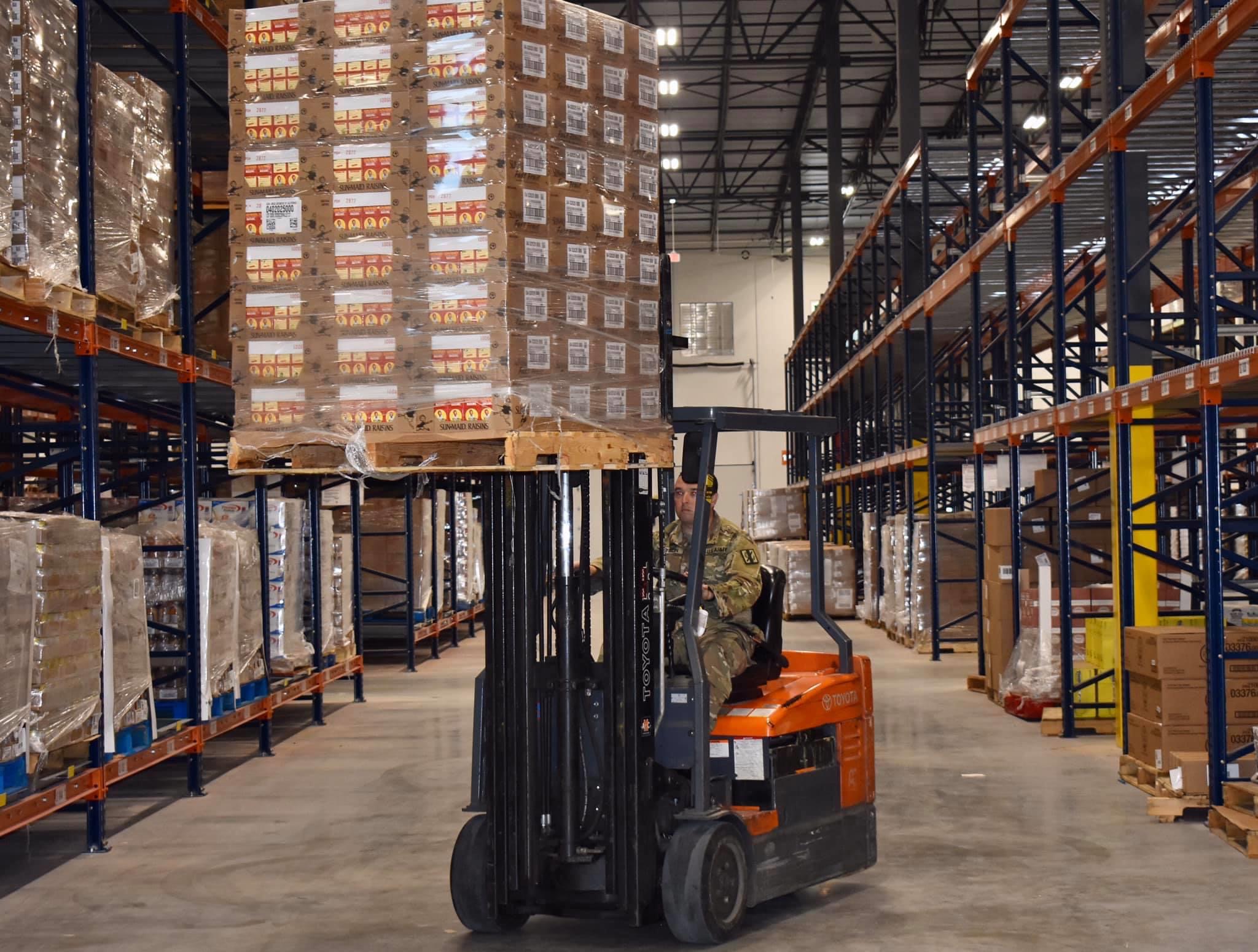 A forklift carries a tall pallet of boxed goods through a large warehouse.