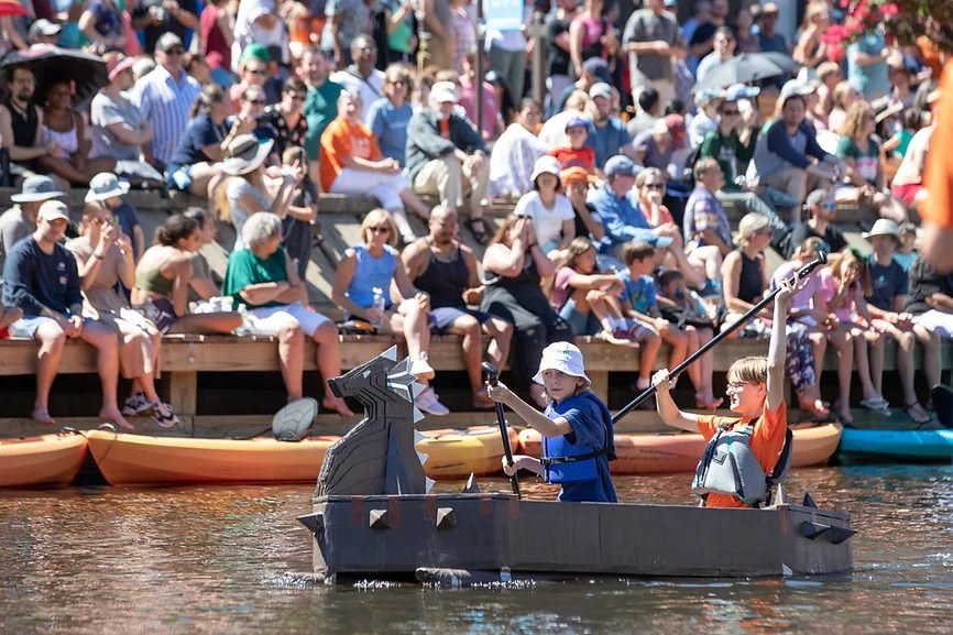 Two people paddling a black dragon-shaped cardboard boat on a lake with spectators on docks.