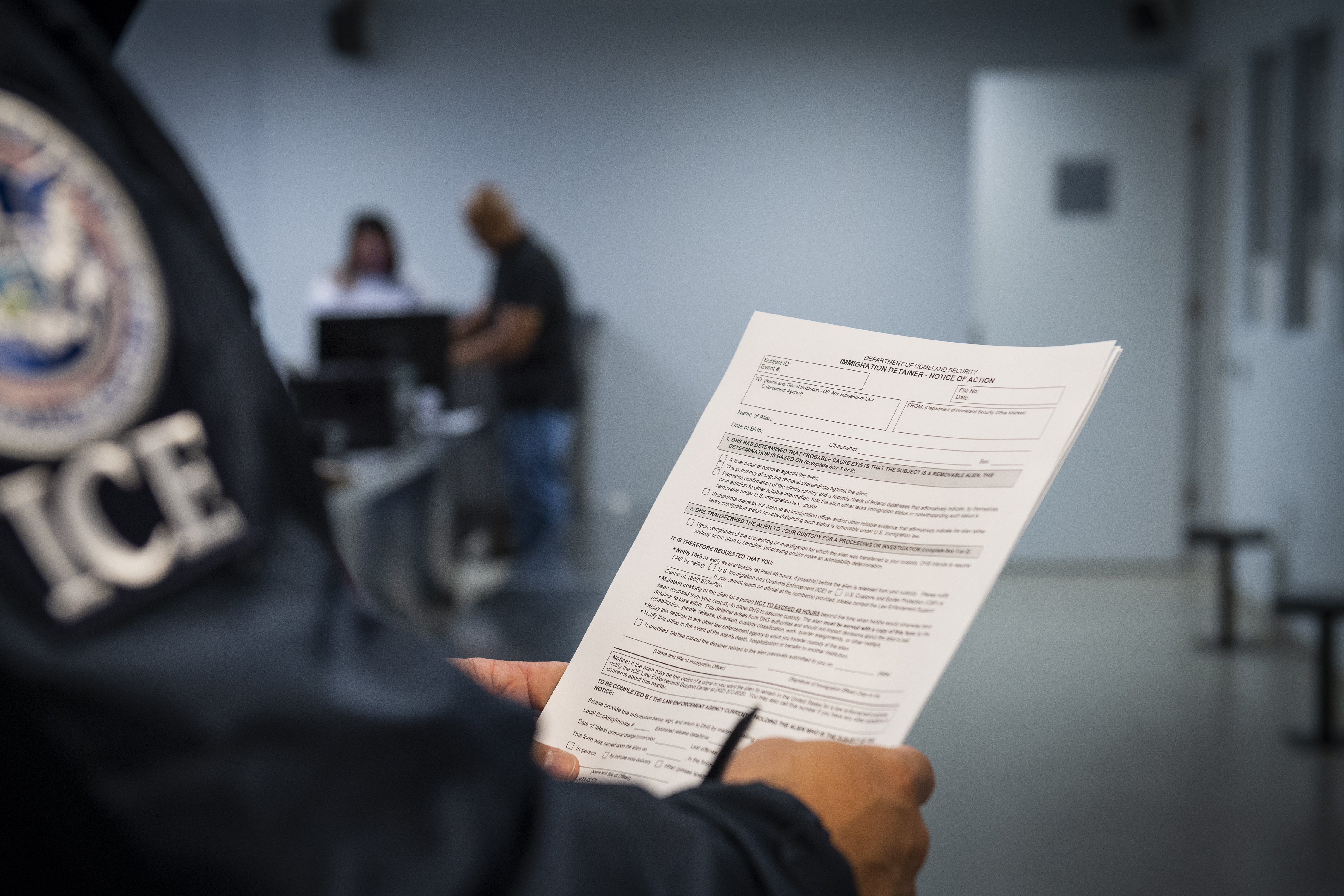 Person wearing an ICE jacket holds a printed immigration detainer form.