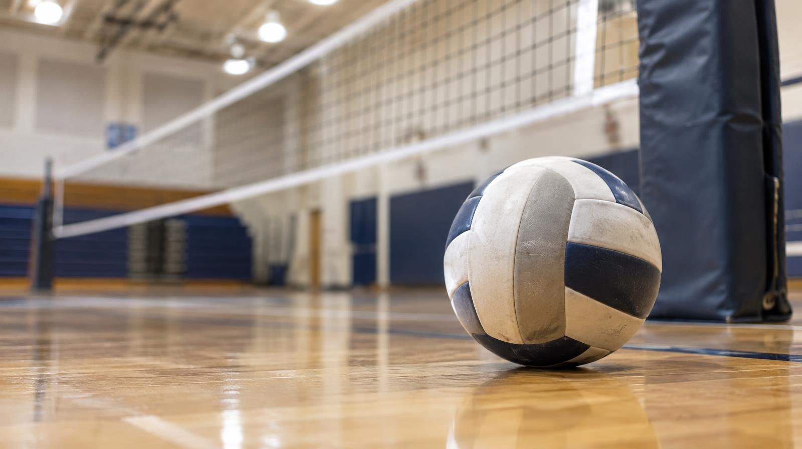A volleyball resting on a hardwood gym floor beside a net post.