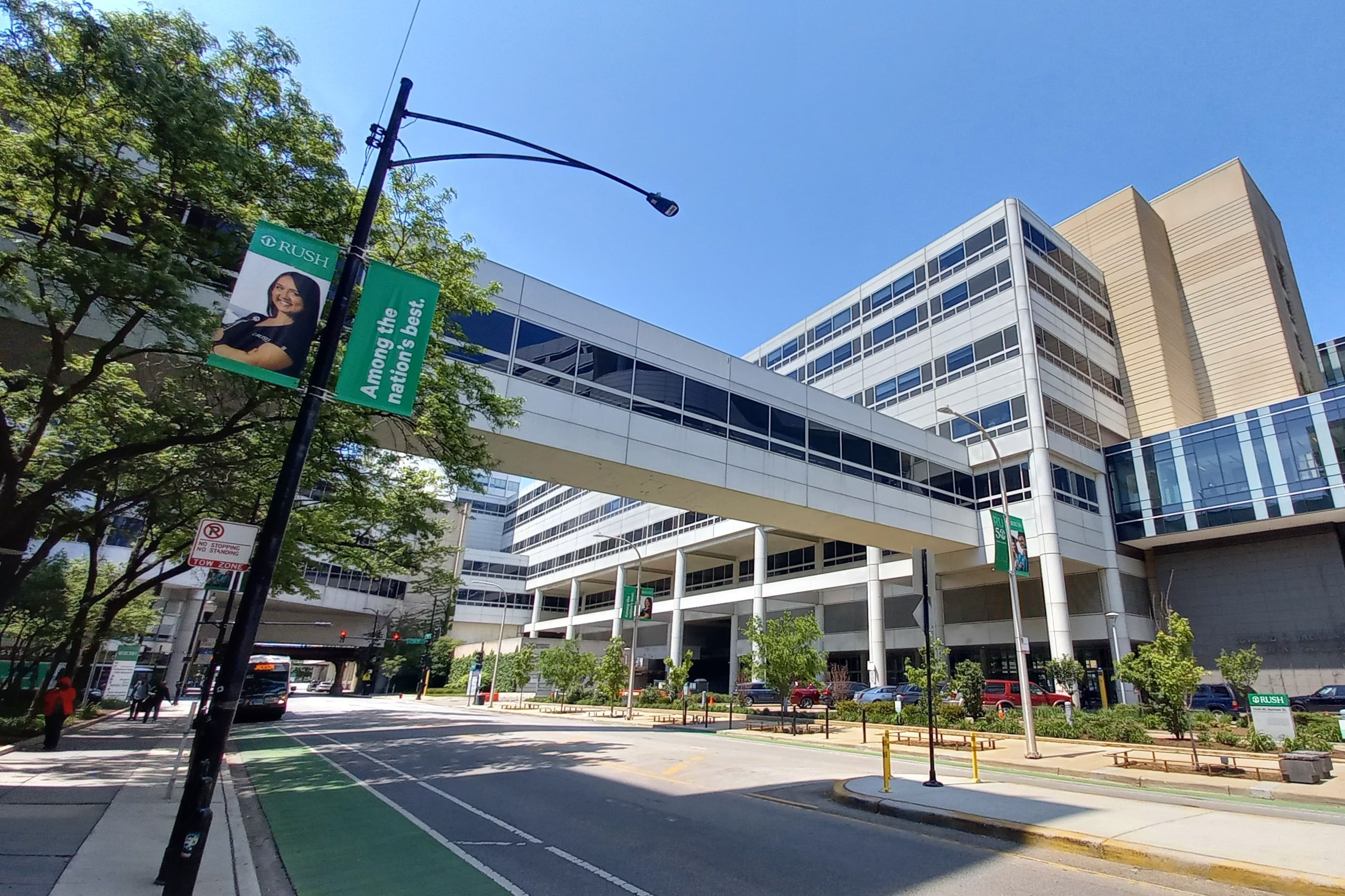 Multi-story hospital complex with a glass pedestrian skybridge over a tree-lined street.