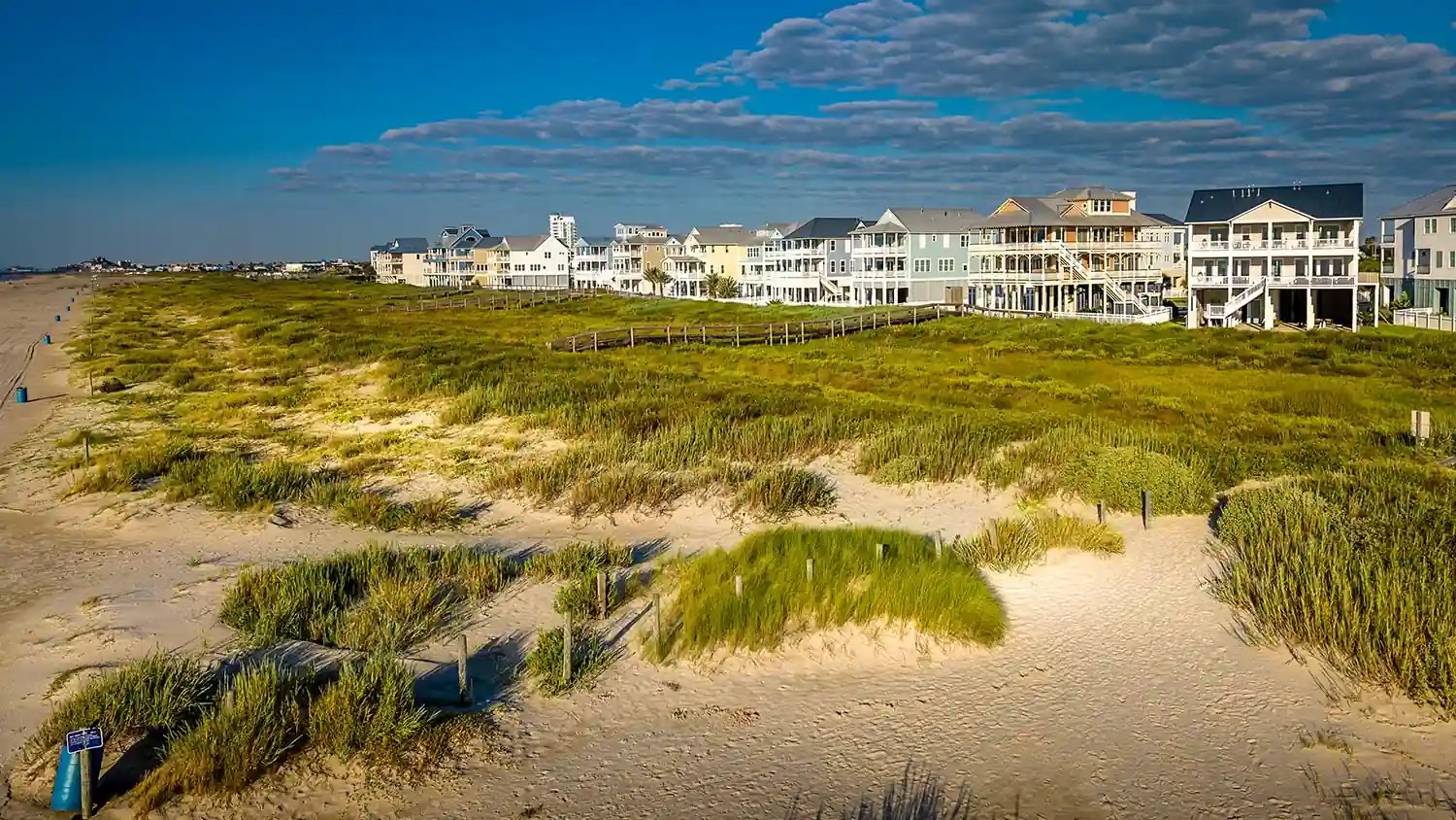 Sandy beach dunes with beach grass and multi-story beachfront homes under a blue sky.