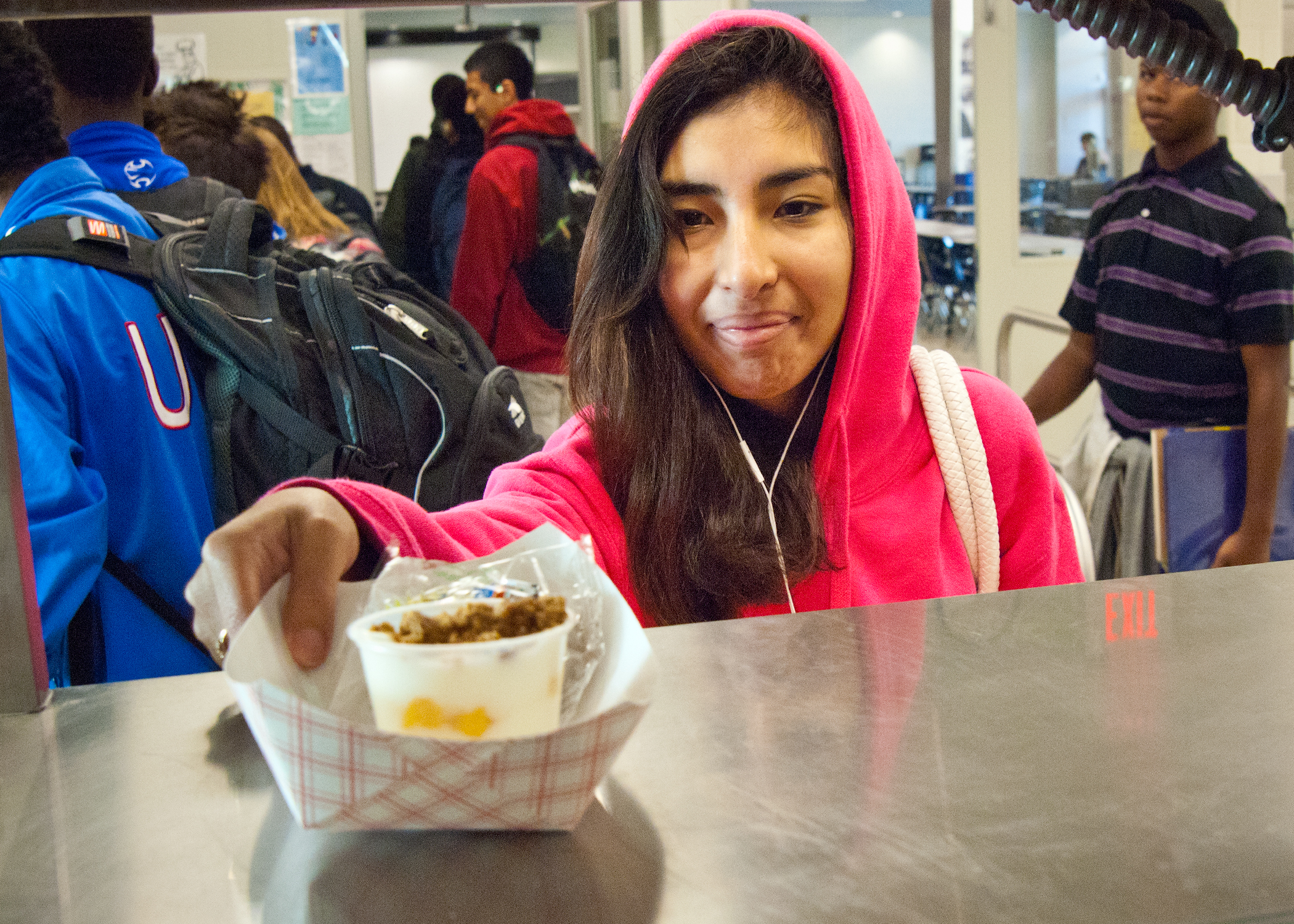 A teenage girl in a pink hoodie reaches for a food tray in a school cafeteria.